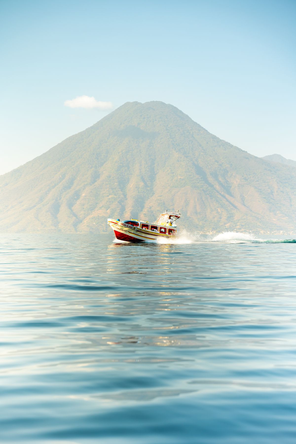 Colorful boat sailing quickly over a calm lake, with a large volcano in the background.