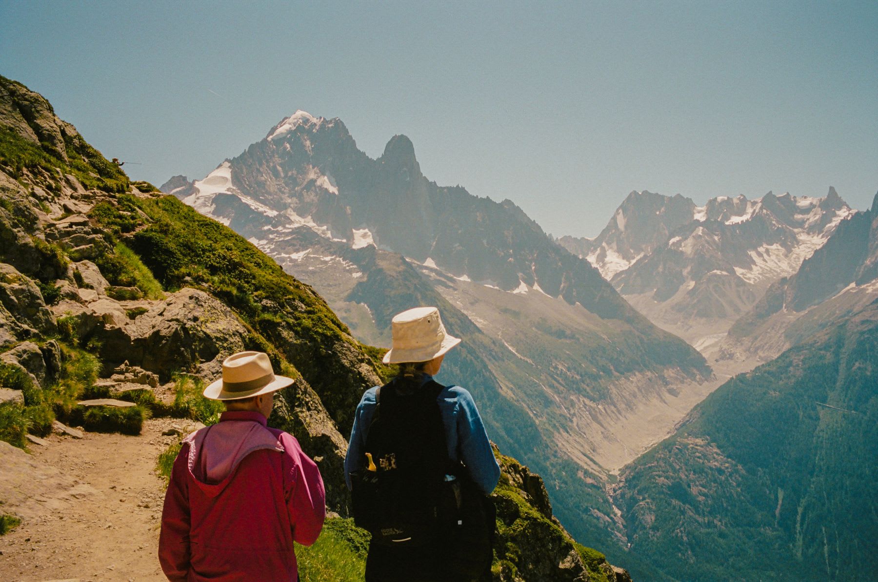 Two hikers in hats stand on a mountain trail, admiring the panoramic view of a snow-capped alpine valley on a sunny day.