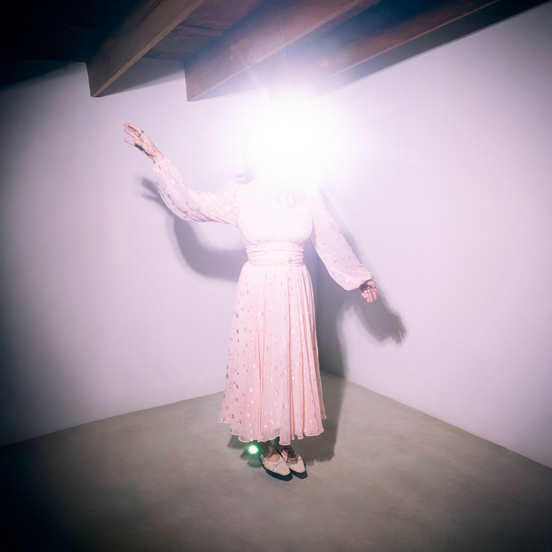 A person in a long, pink, polka-dotted dress stands in a corner of a room with wooden beams on the ceiling, with a bright light obscuring the face.