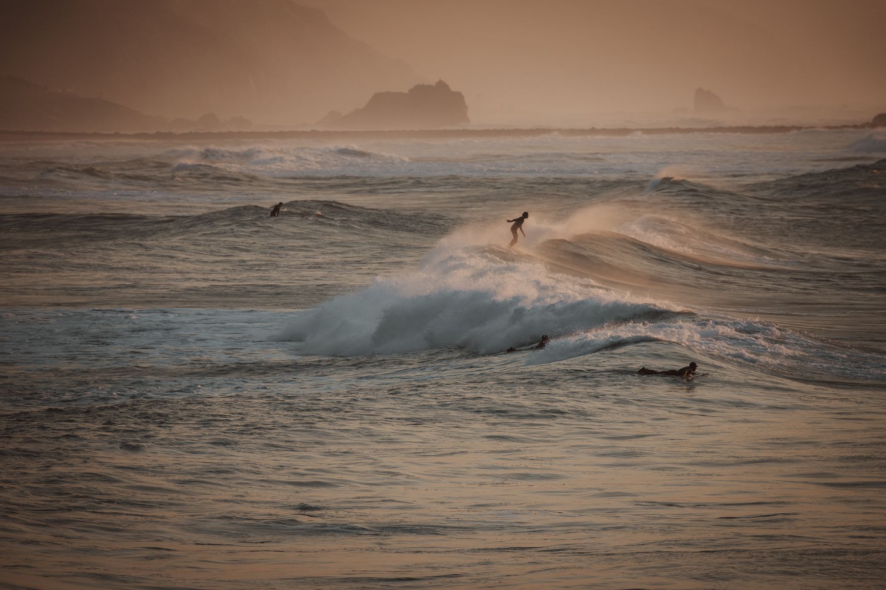 Four surfers in the sea, with three of them trying to catch waves and one riding a wave.