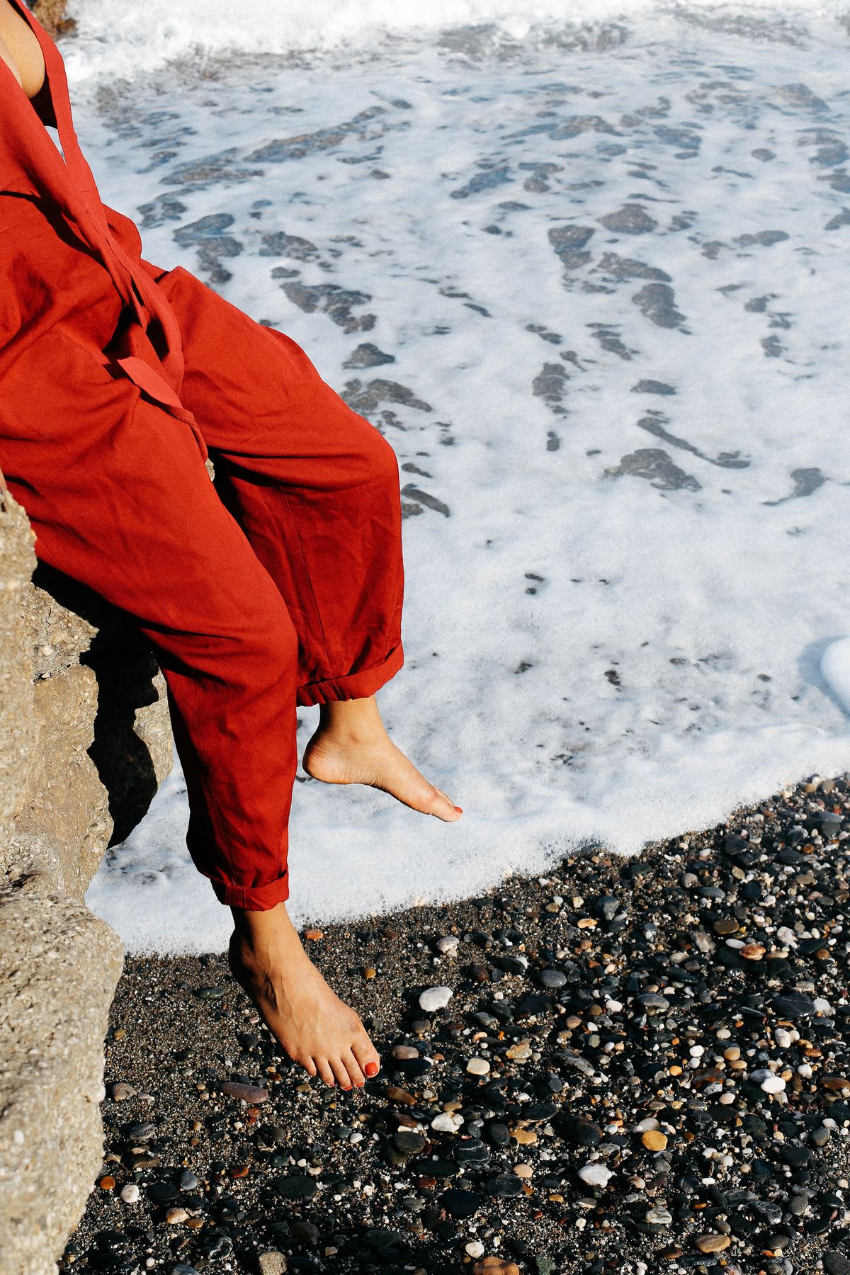 A woman models a red jumpsuit while sitting on a boulder on an ocean coast.