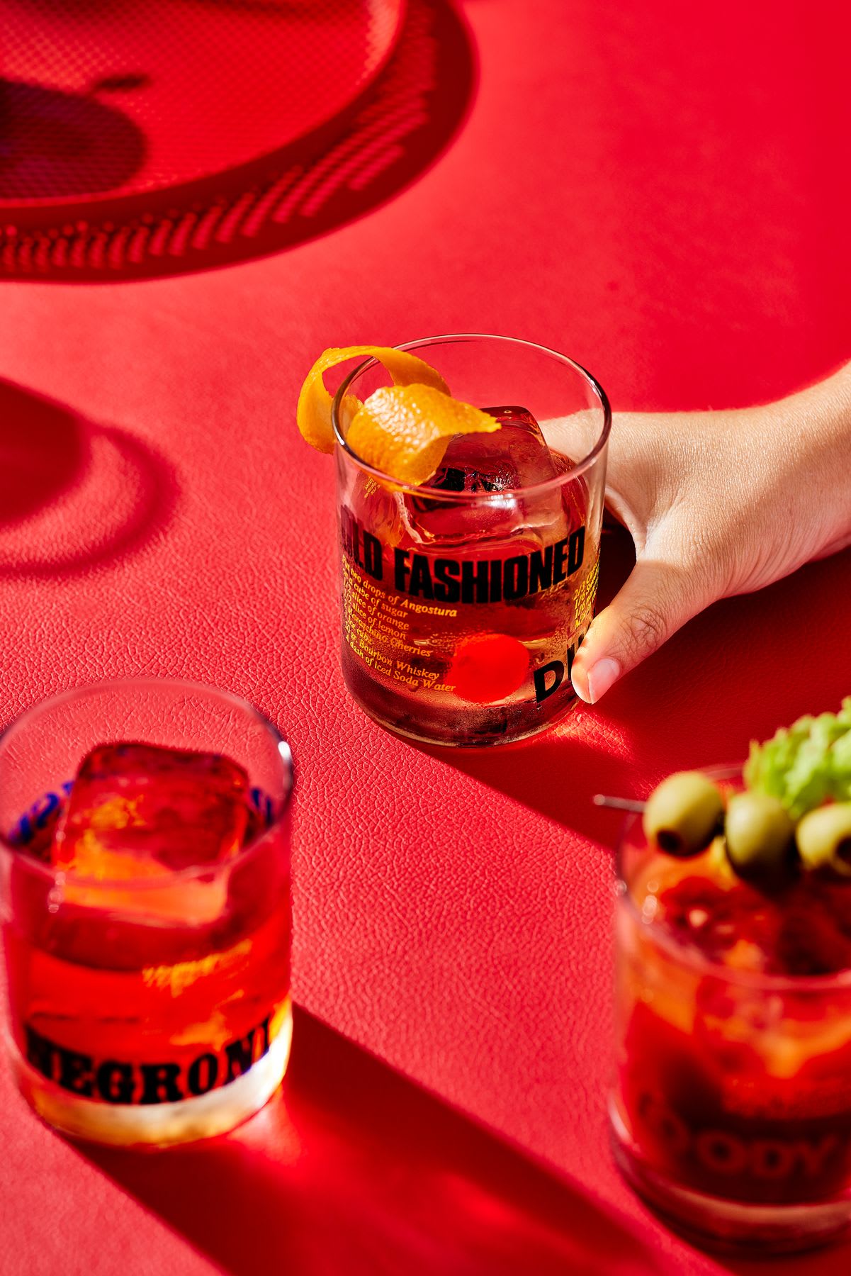 Three cocktails sit on a bright red surface: one ‘Old Fashioned’ with an orange peel and cherry, one ‘Negroni’ with ice, and a third garnished with olives, all with vibrant colors and a hand holding the Old Fashioned.