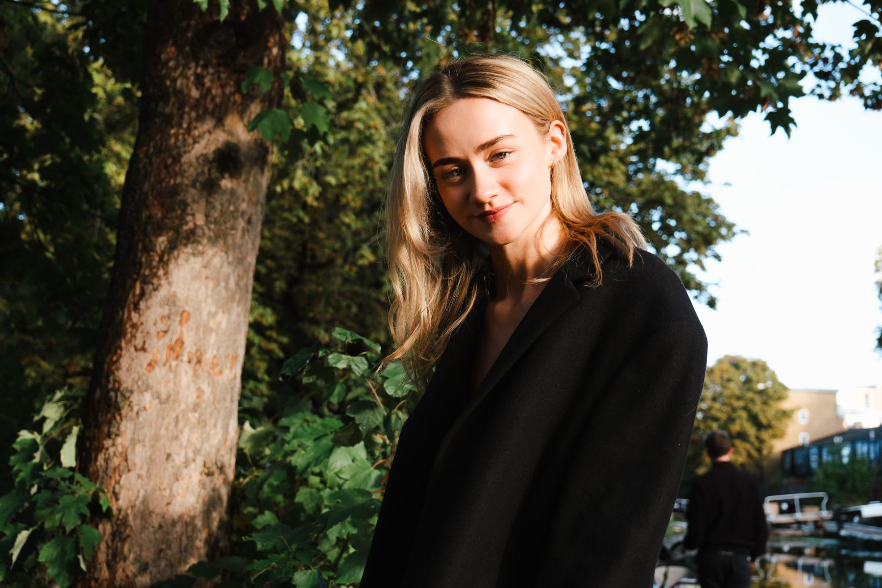 A young woman with long blonde hair smiles gently while wearing a black coat and standing outdoors next to a tree.