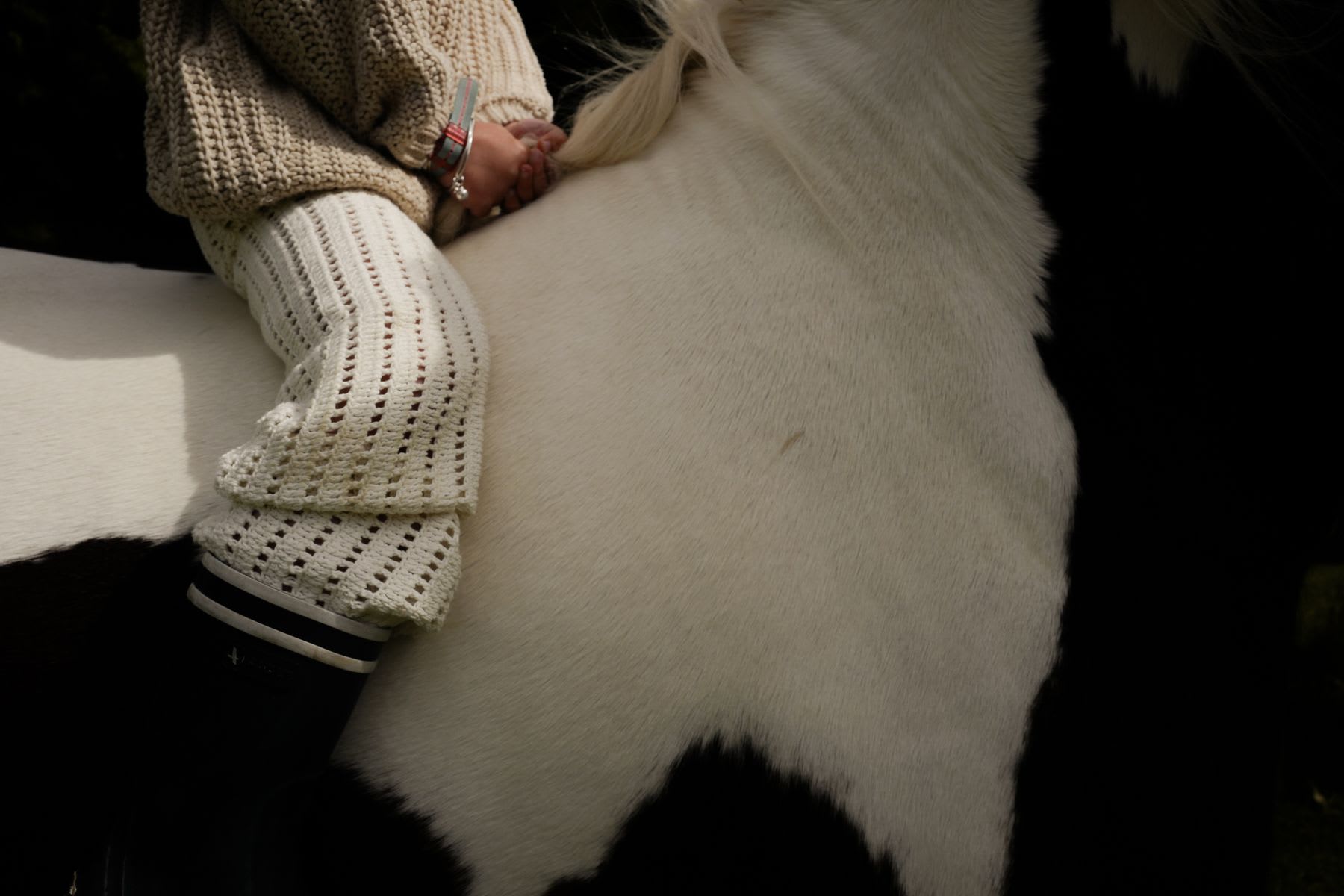 A child wearing a cozy beige knit sweater and white leggings holds onto the mane of a white and black horse.