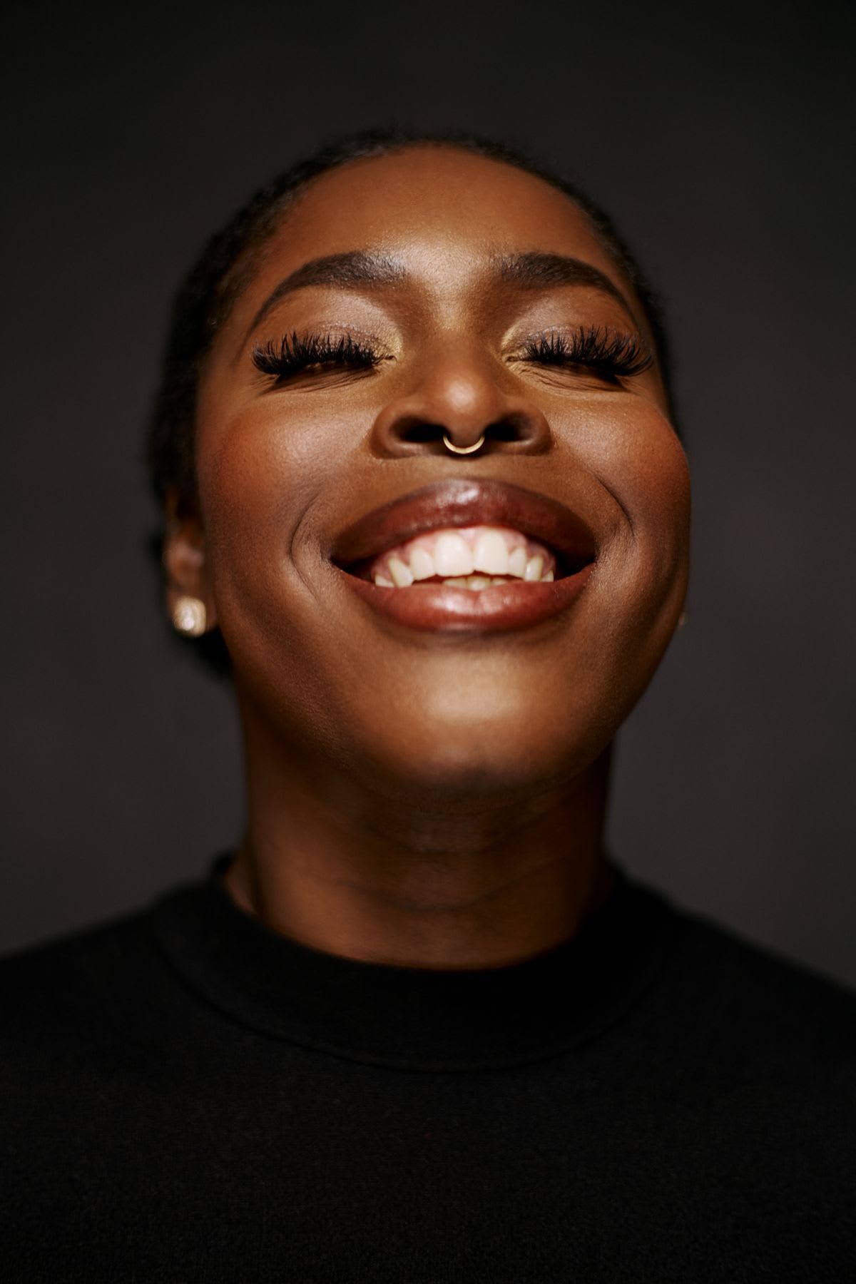A person smiling, with make-up and a nose ring, against a dark background.