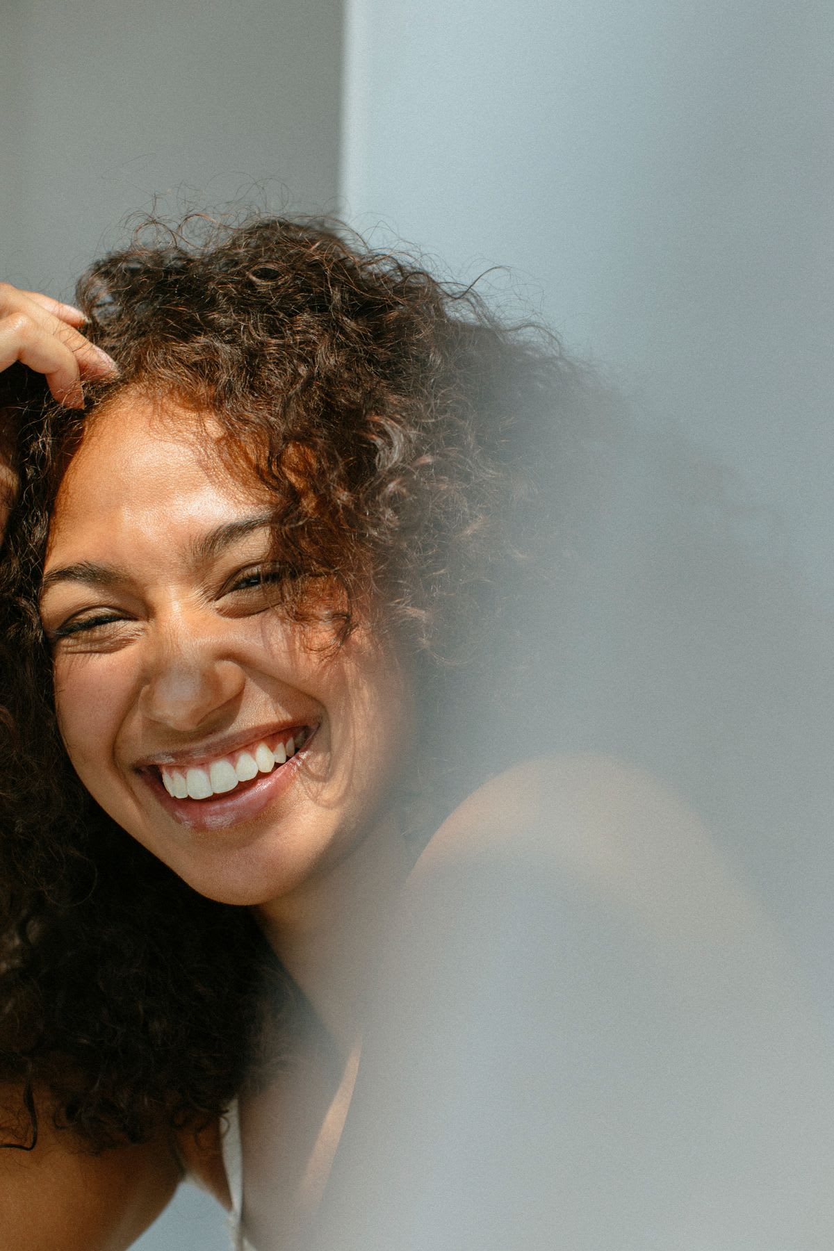A curly-haired woman beams with genuine joy in a softly lit close-up, her bright smile and sparkling eyes popping against a subtle, out-of-focus foreground.