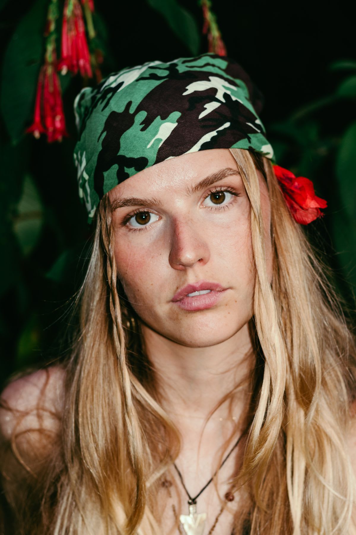 A close-up portrait of a young blonde woman wearing a camouflage headband and a red flower in her hair against a dark leafy background.