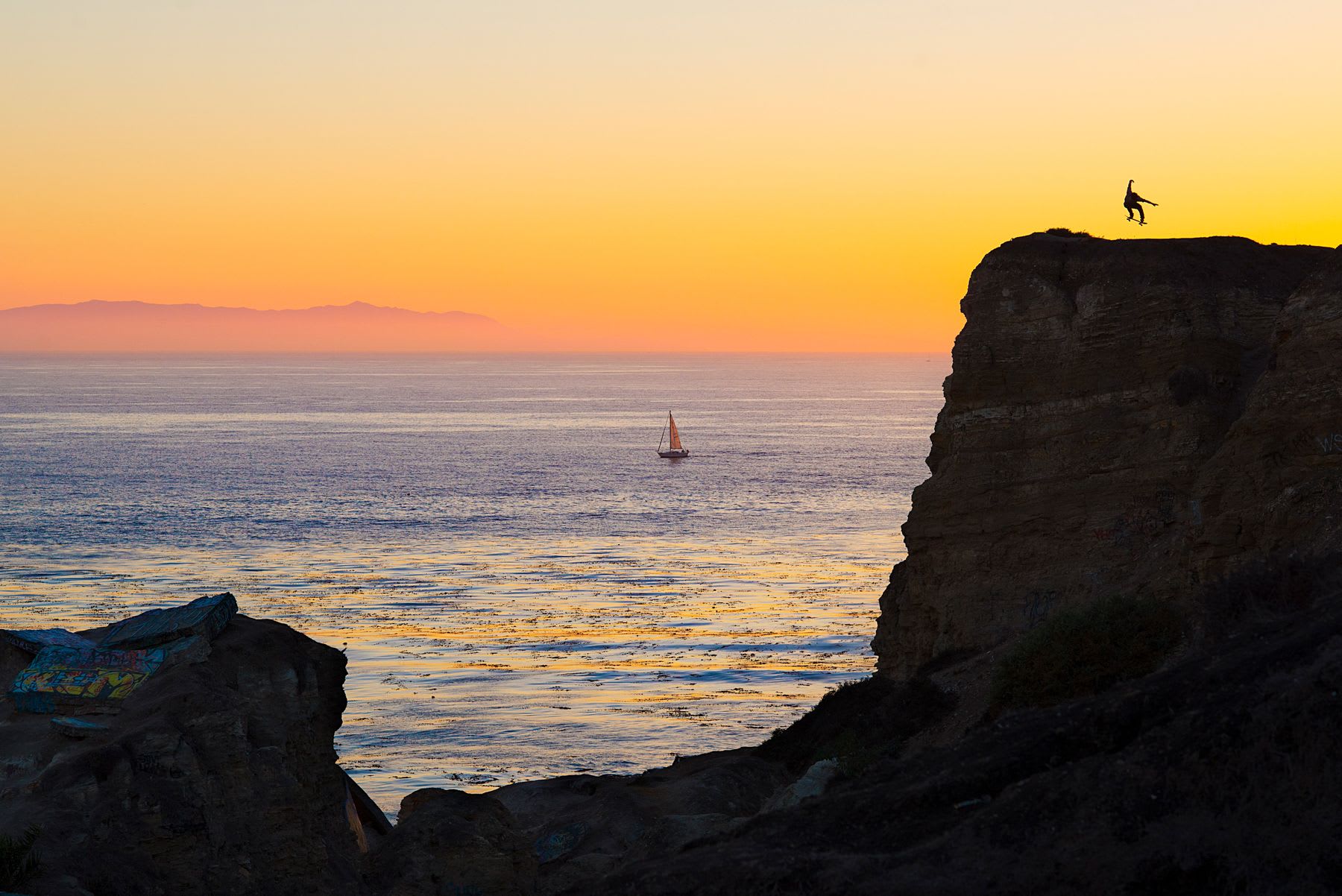 A man performs a trick with a skateboard on a cliff overlooking an ocean, with a small boat sailing at sunset