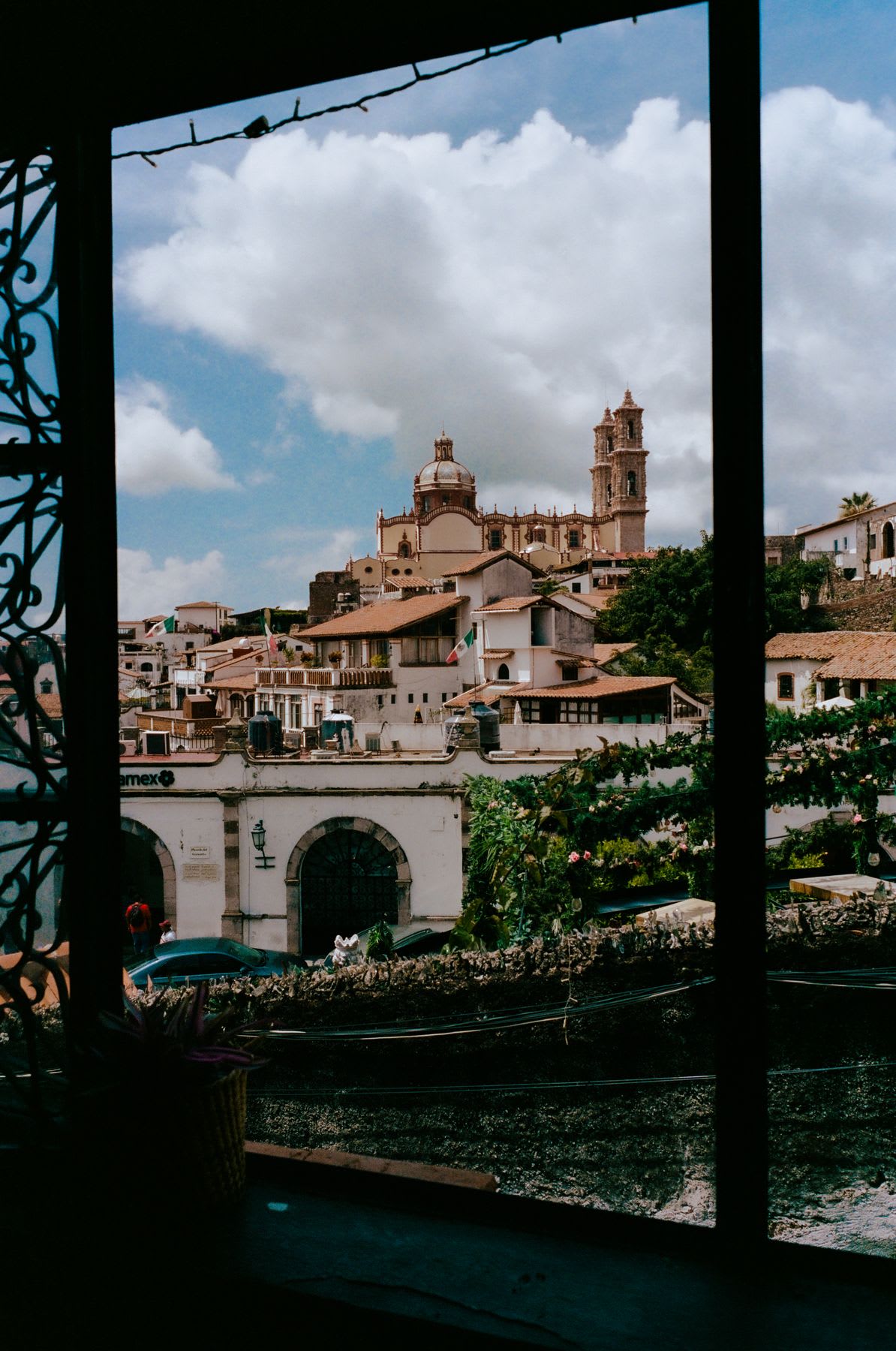 Scenic view through a window frame of a small town with whitewashed buildings, a large church with a dome, and a tall tower in the background. The sky is partly cloudy, and green plants and Mexican flags are visible in the foreground.