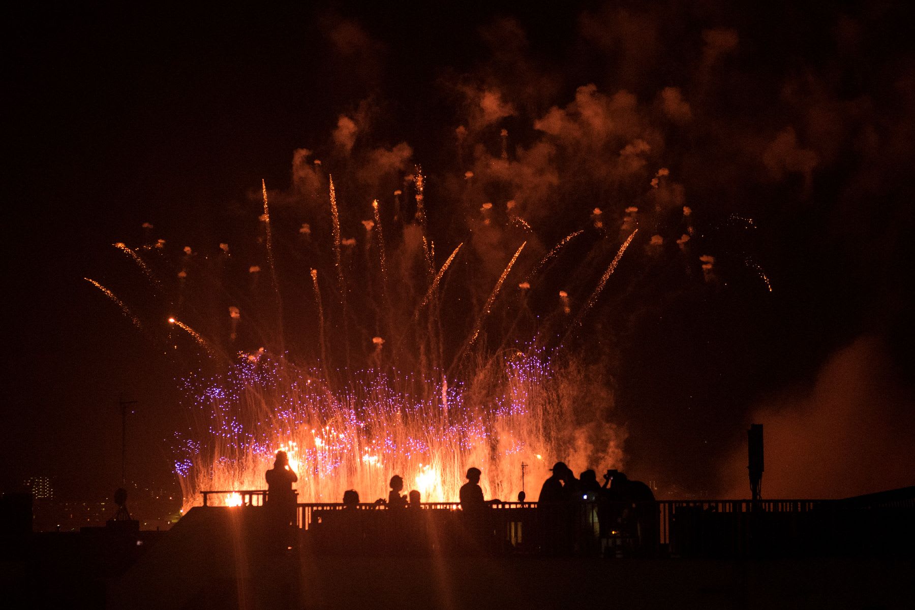 People gather at the edge of a bridge, celebrating with fireworks illuminating the night sky.