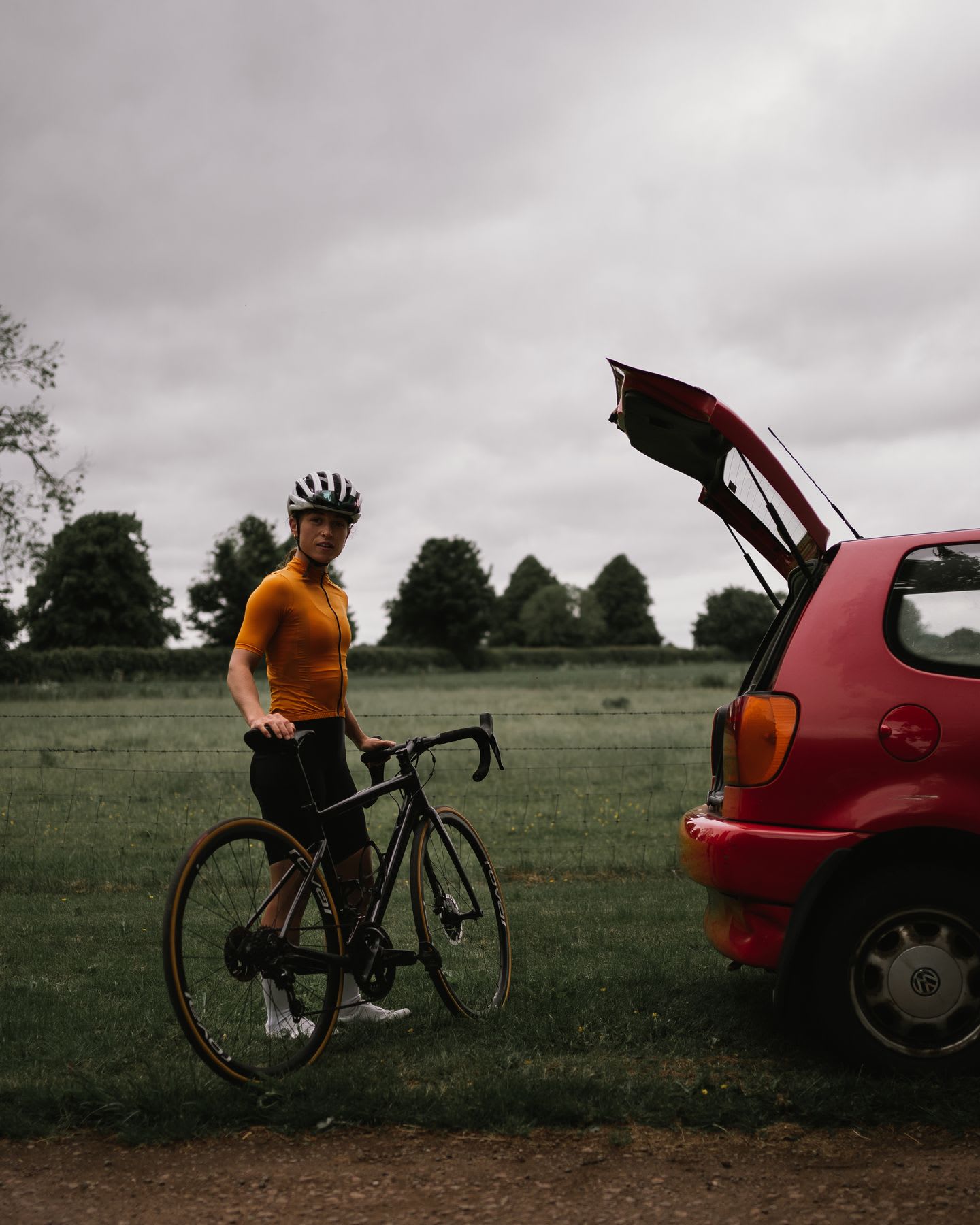 A biker in a white helmet pauses by her bike and a red car with its trunk open, on a grassy field under cloudy skies.
