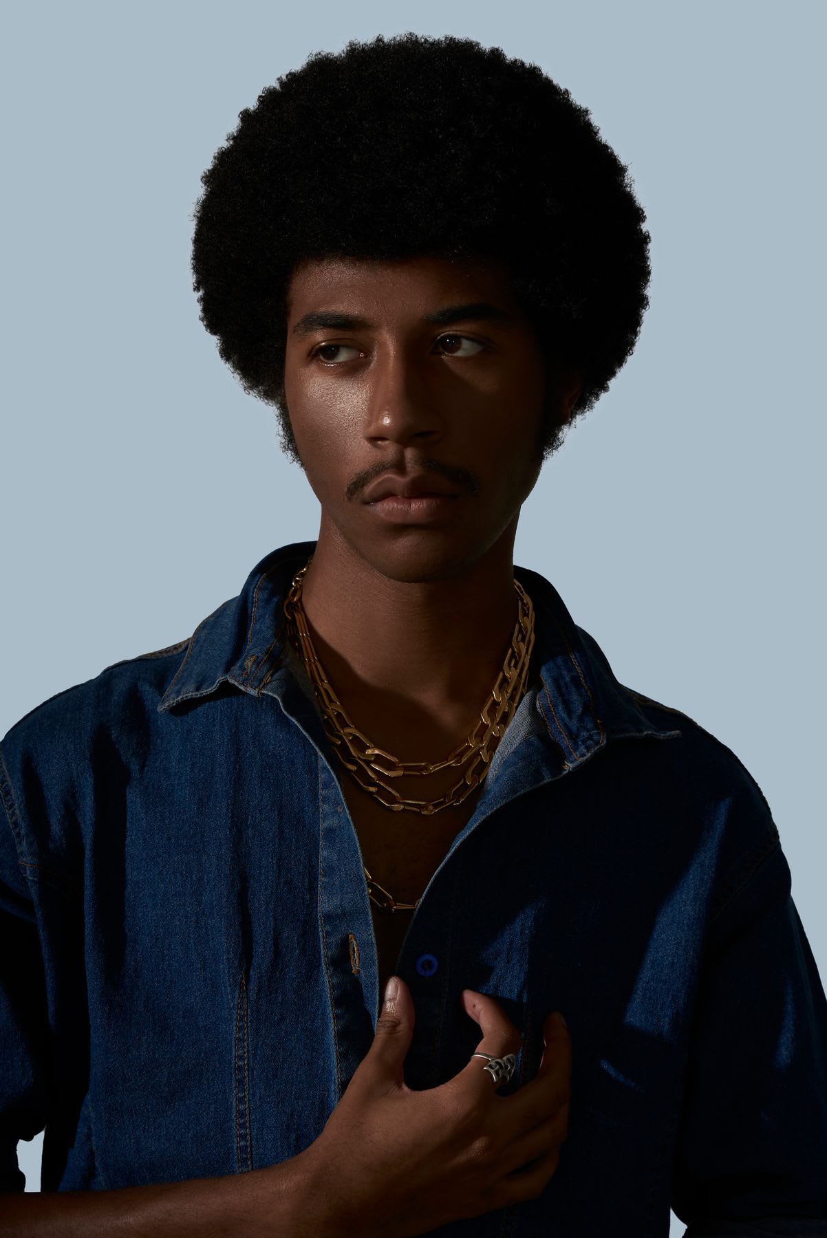 Young man with an afro hairstyle, wearing a denim shirt and gold chains, looking off to the side against a plain background.