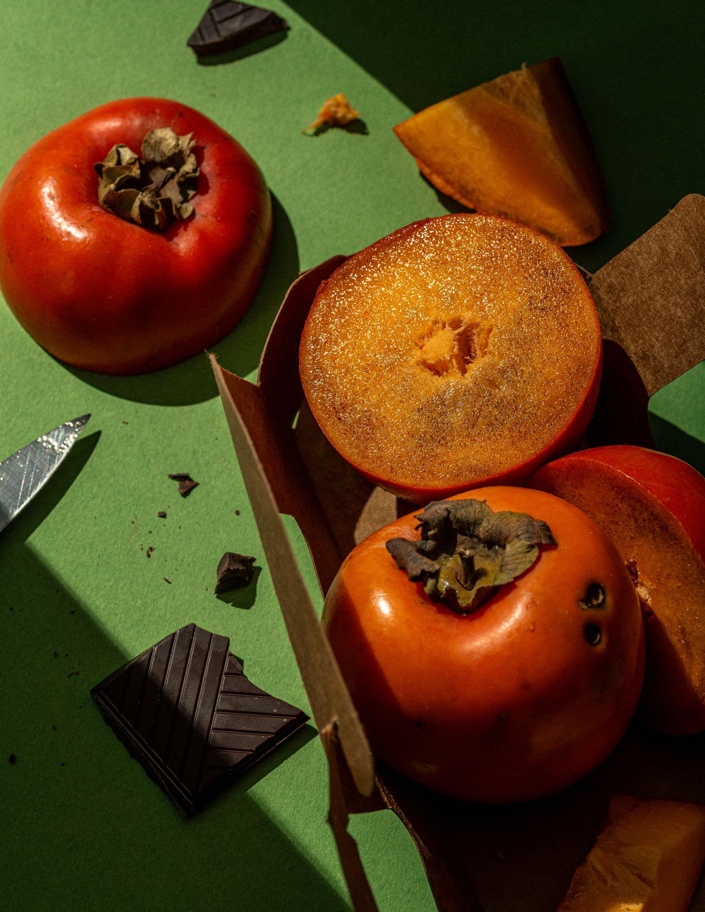 A vibrant still life captures persimmons in various states whole, halved, and sliced arranged on a green surface alongside pieces of dark chocolate and a knife, all bathed in a strong, directional light that casts distinct shadows.