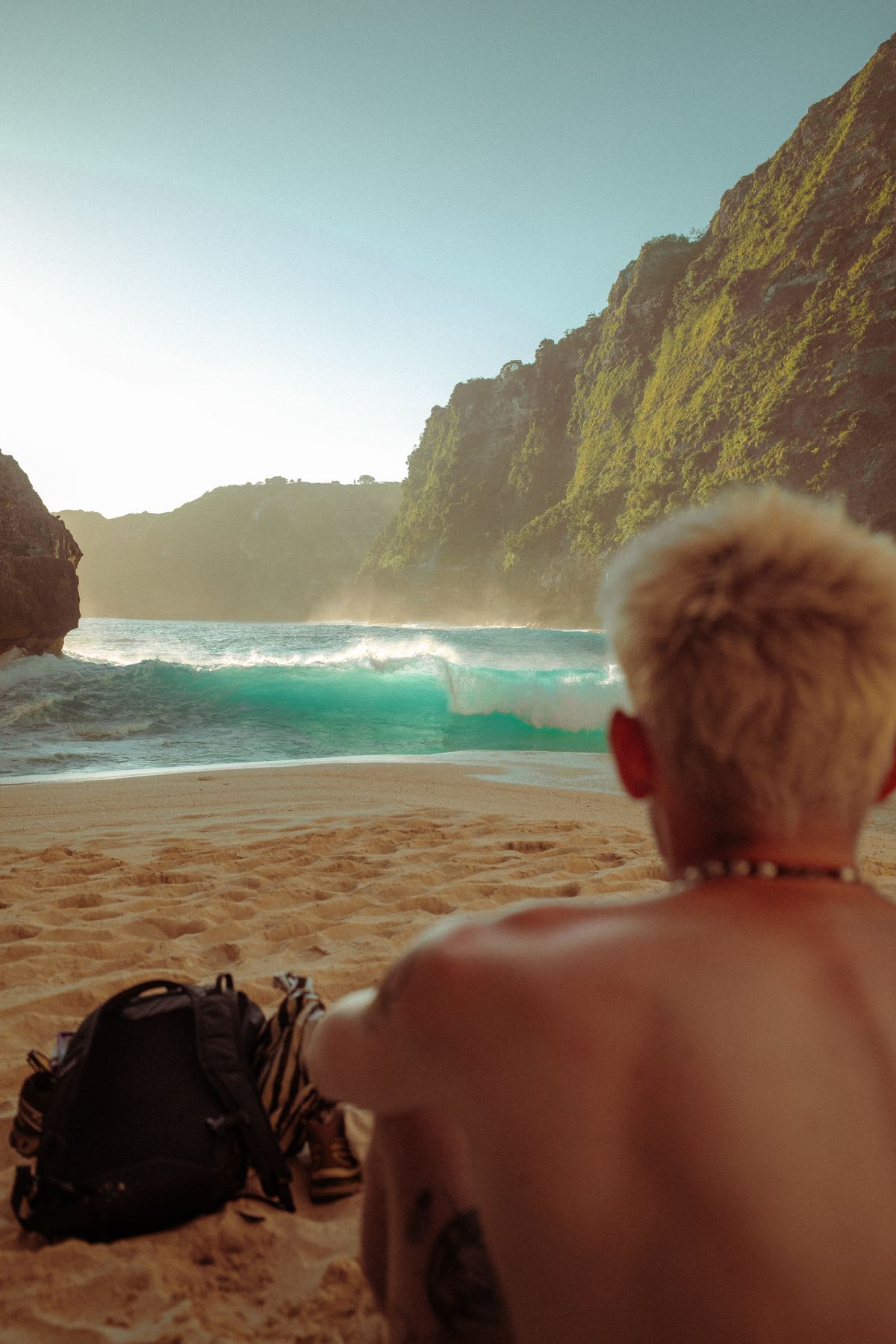 From a back view, a shirtless person with blond hair sits on a sandy beach and watches the turquoise waves crash against the shore.