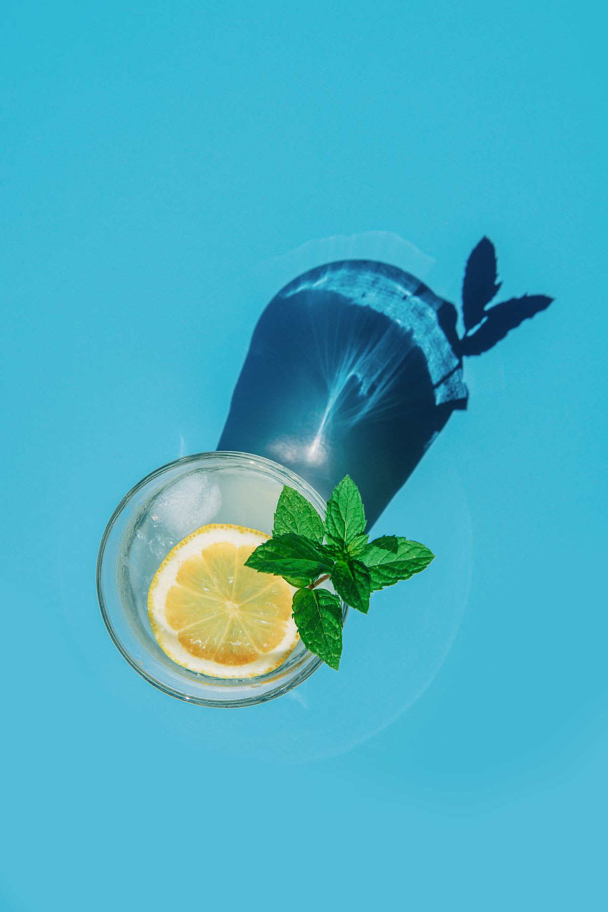 Drink with ice, a slice of lemon, and fresh mint leaves, casting a shadow on a blue background.