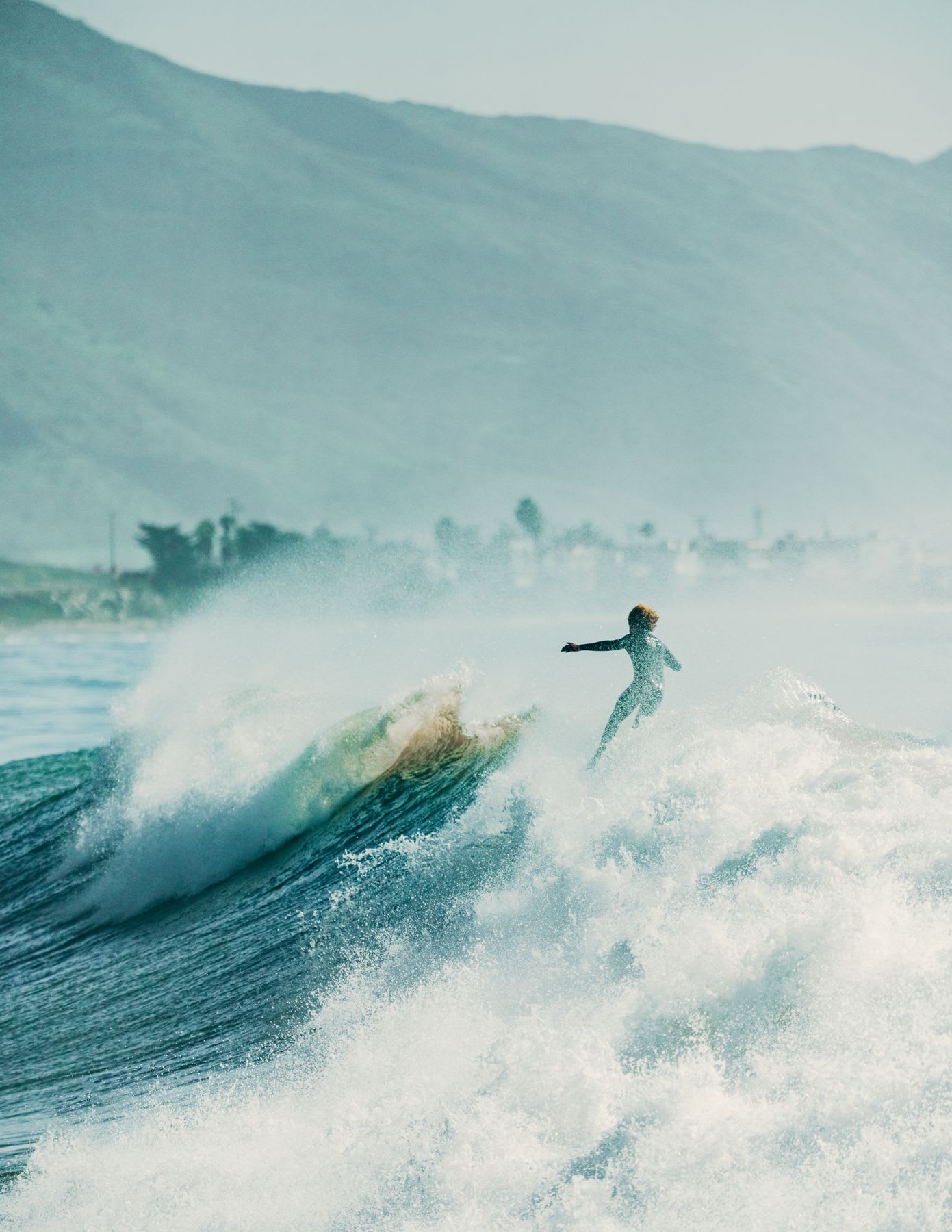 A lone surfer carves down a large, splashing ocean wave with a hazy mountainous coastline in the background.