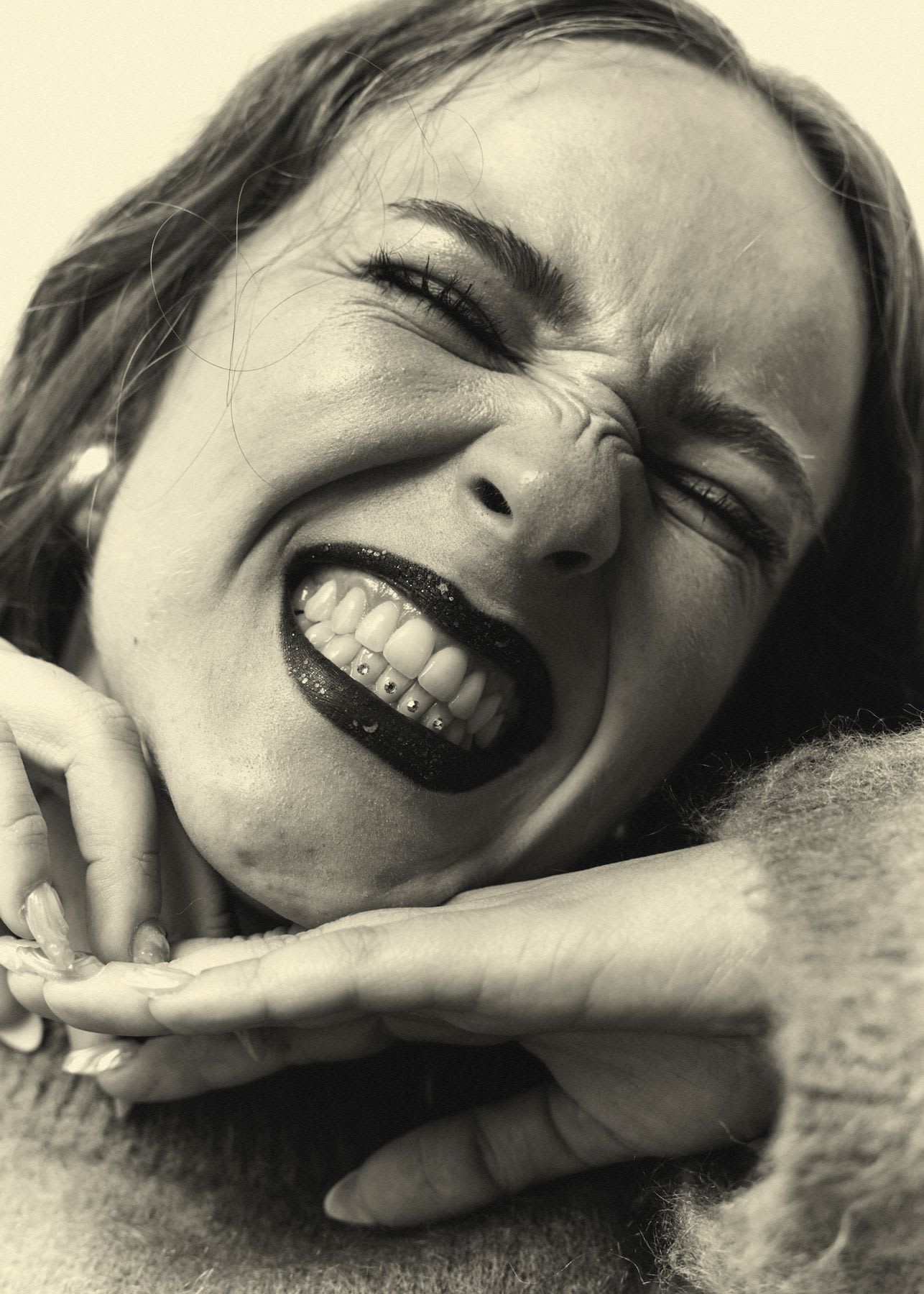 A joyful woman with dark lipstick and jeweled teeth scrunches her face in a big smile in this close-up, black and white portrait.