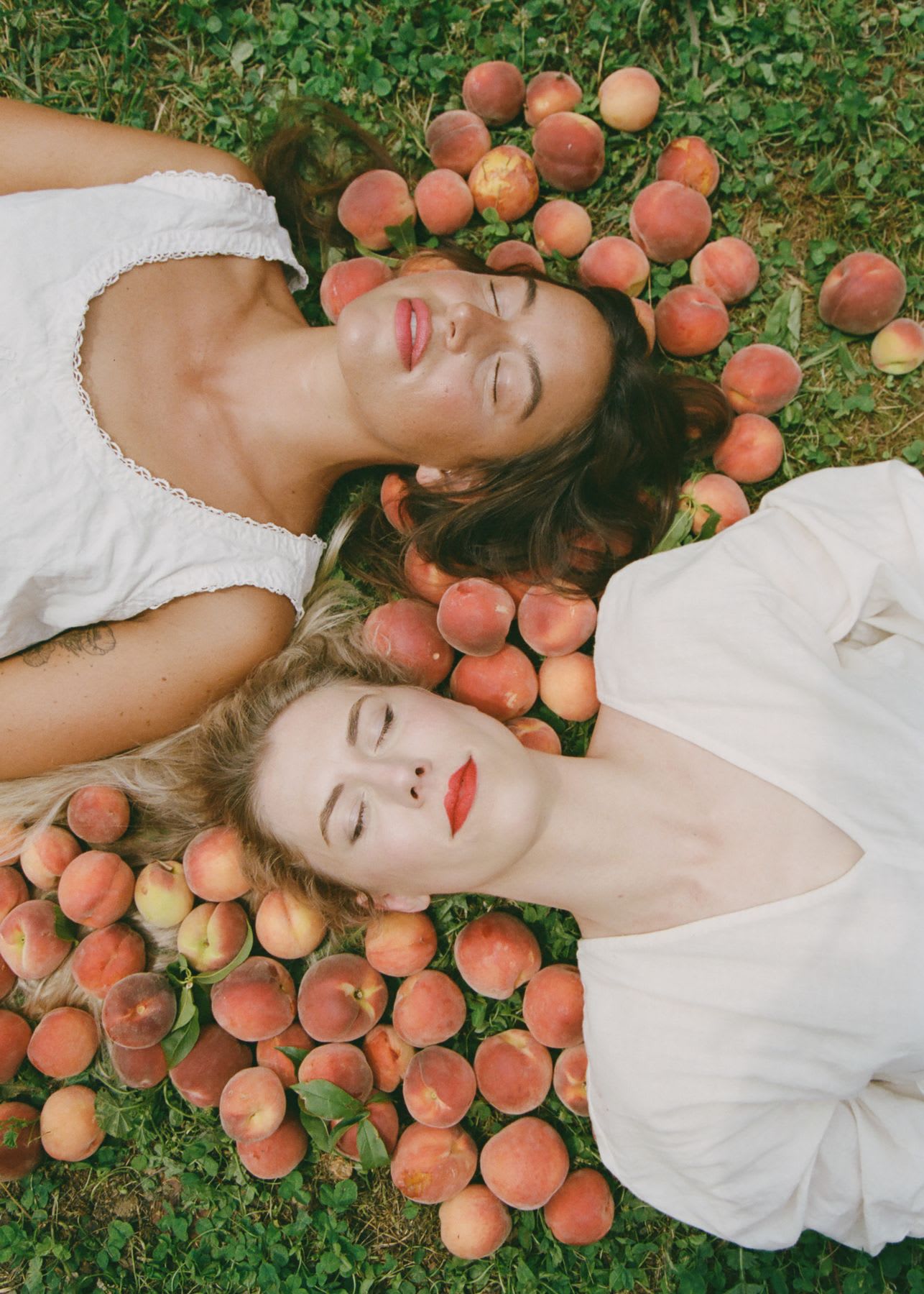 Two women with closed eyes lie on green grass, surrounded by freshly picked peaches.