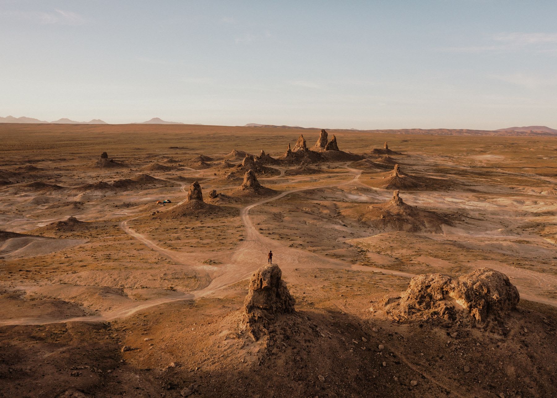 A lone person stands on top of a rock pinnacle, looking out over a vast and arid desert landscape with unique geological formations.