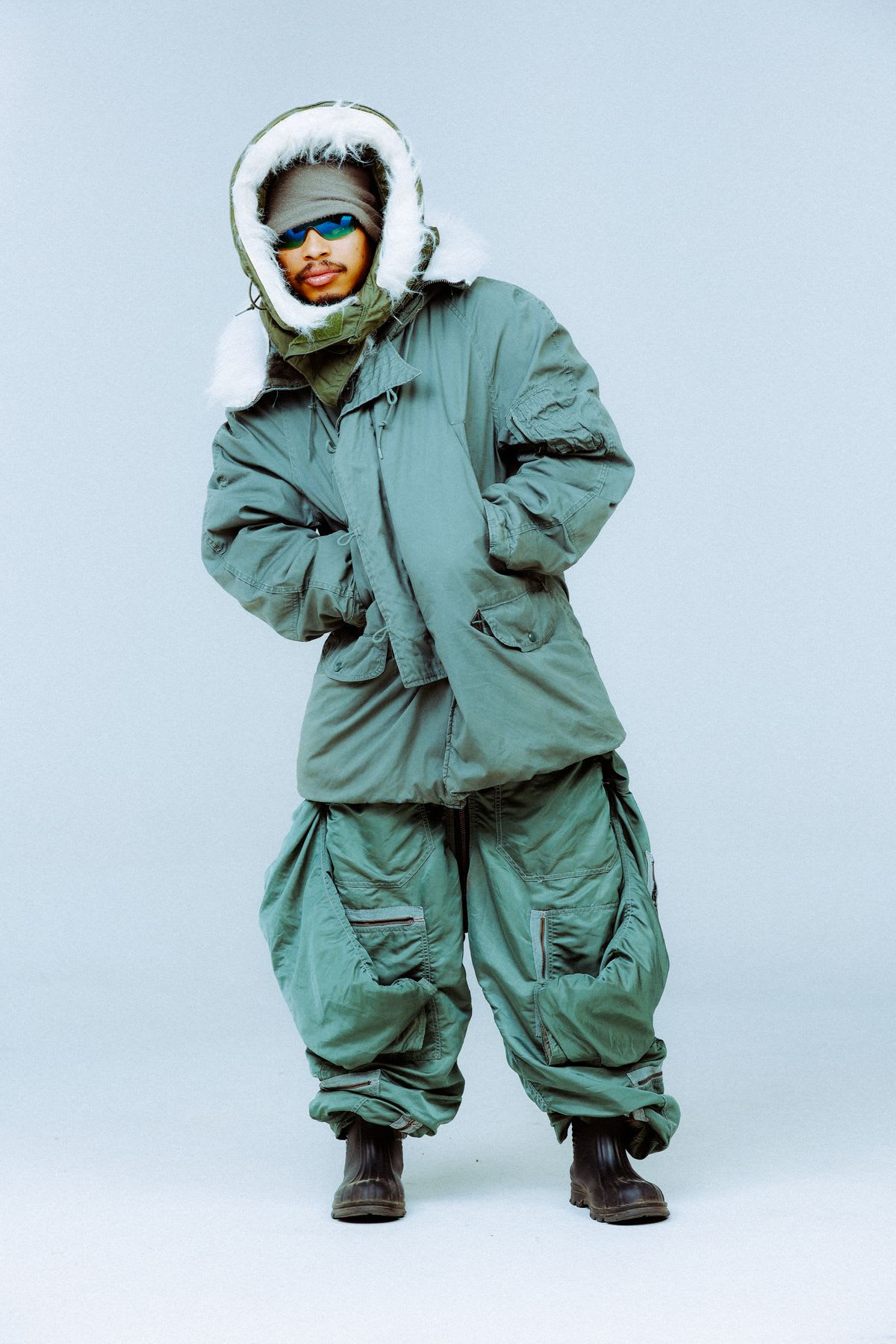 A man poses while modeling a puffy green winter coat as well as very baggy winter pants.