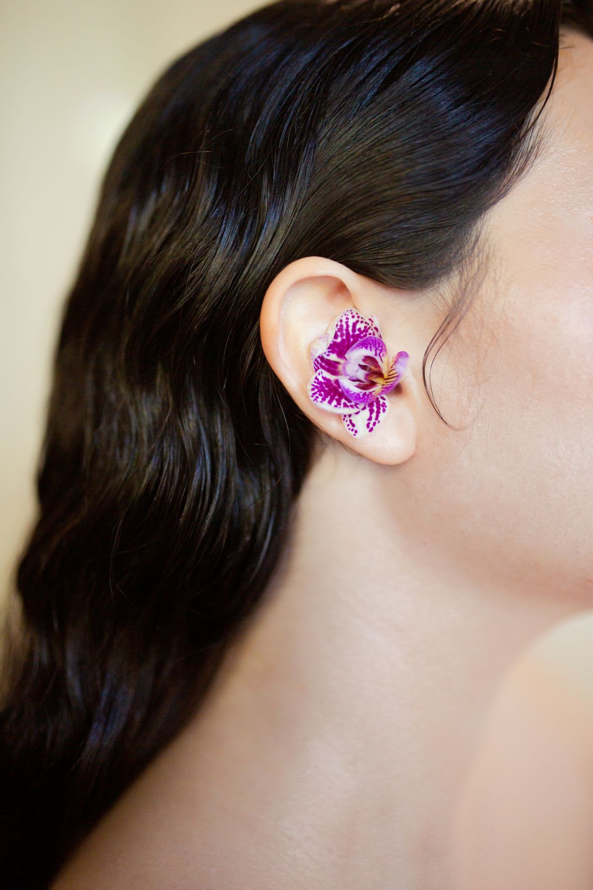 Close-up of a woman’s ear with a purple orchid flower placed inside, surrounded by smooth dark hair.