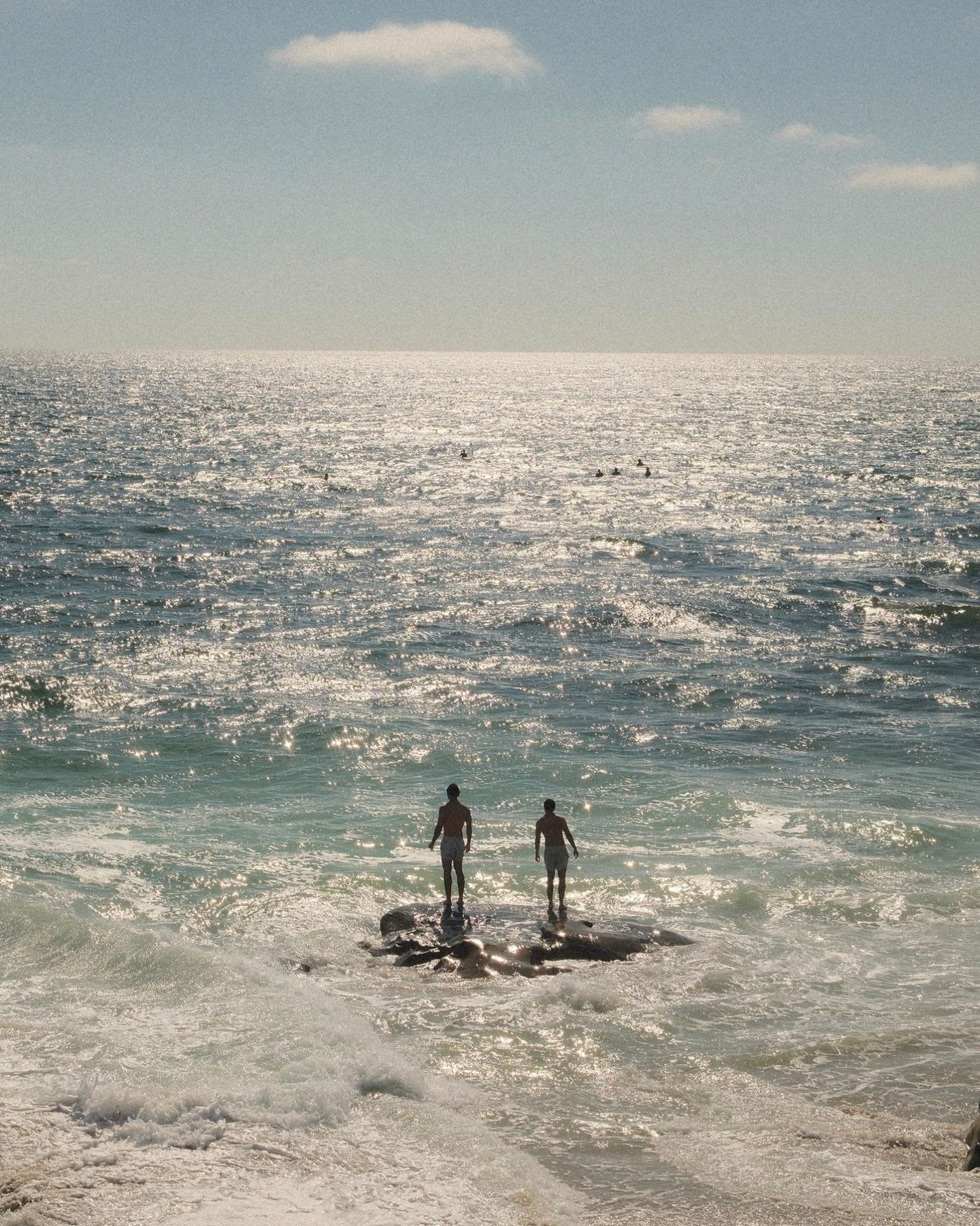 Two men in swim shorts stand on a rock surrounded by waves as they gaze out at the vast, sun-drenched ocean.