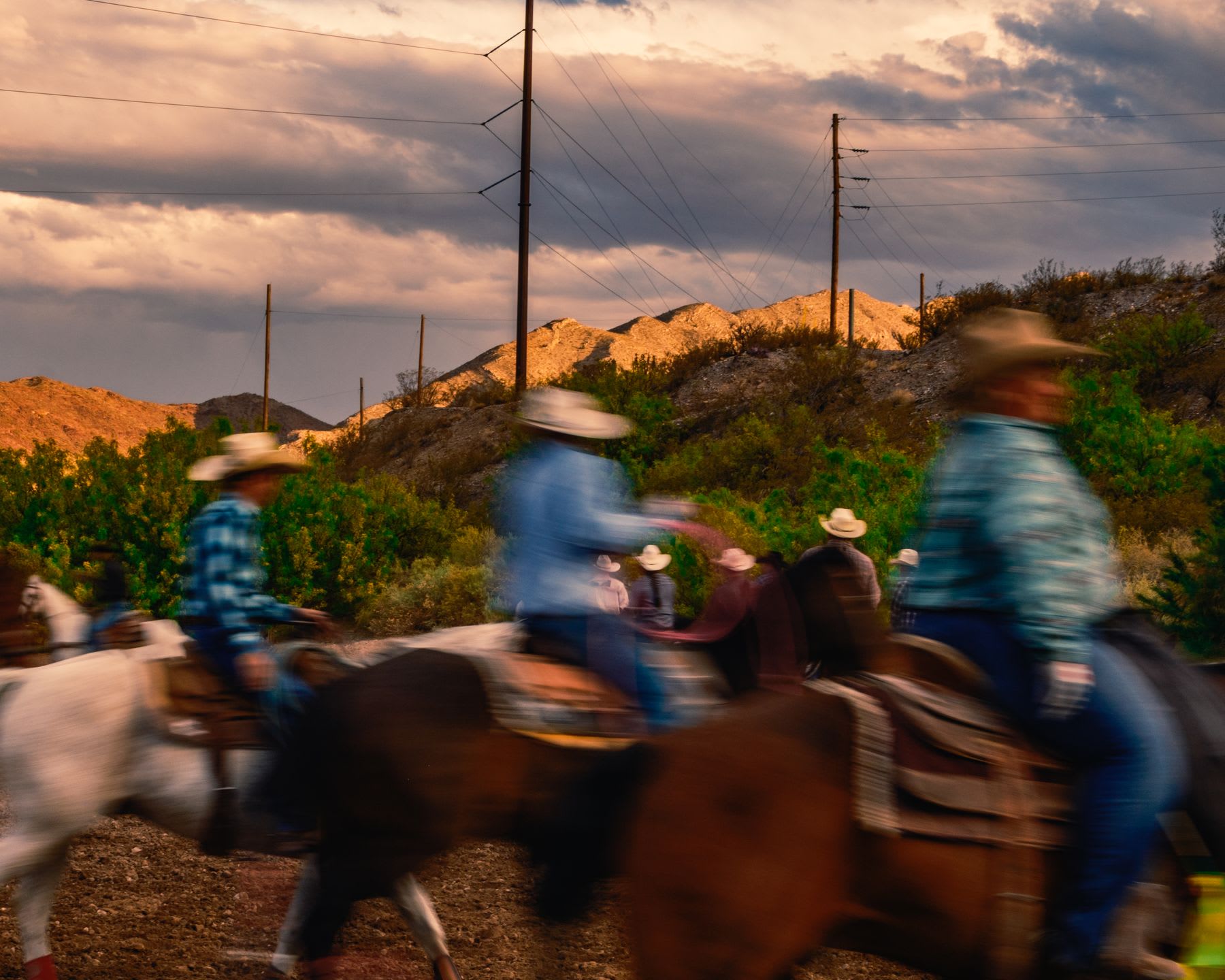 A group of people in western hats are horseback riding through a rugged desert landscape at sunset, captured with motion blur.