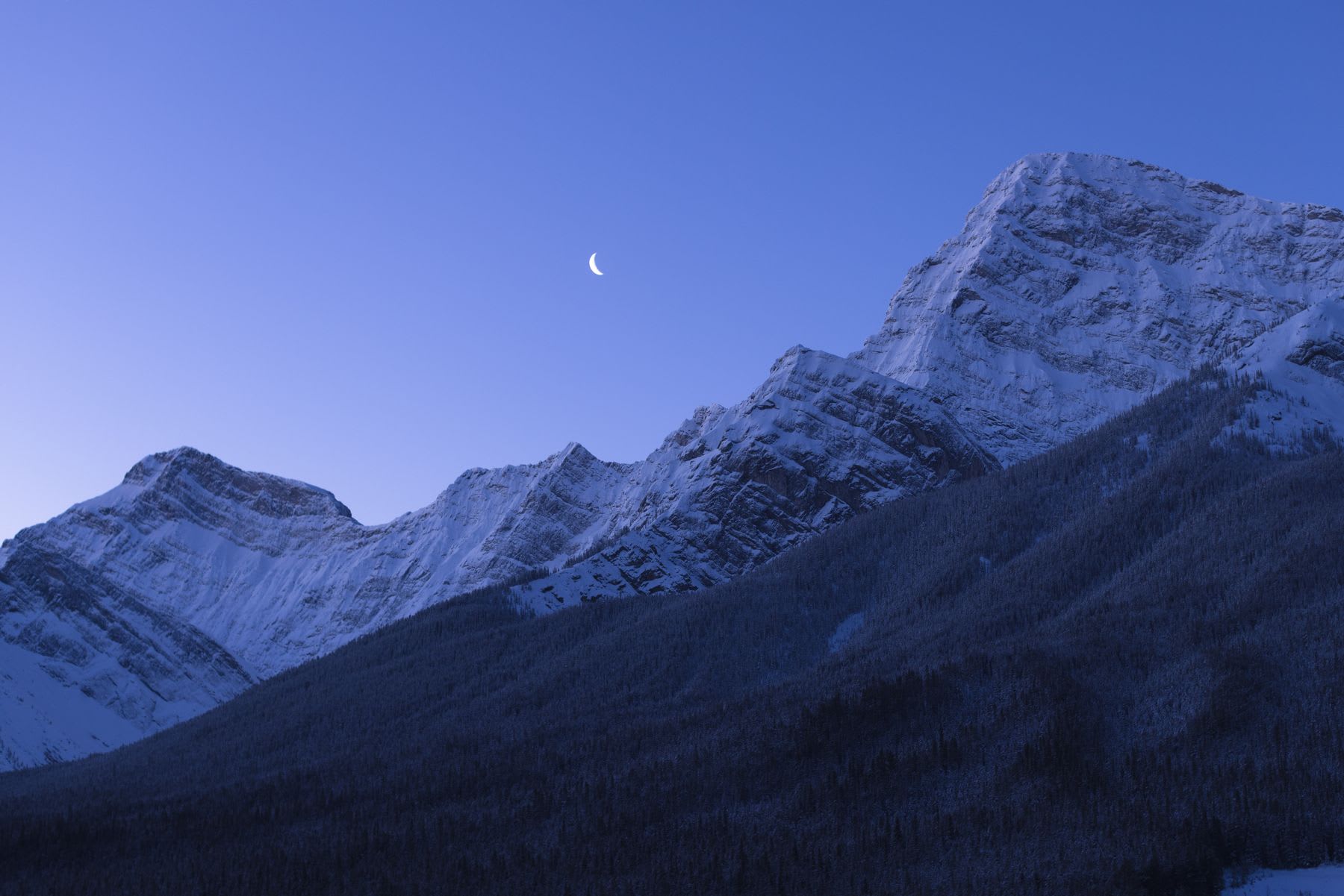 Snow-capped mountains under a blue sky at dusk, with a crescent moon visible.