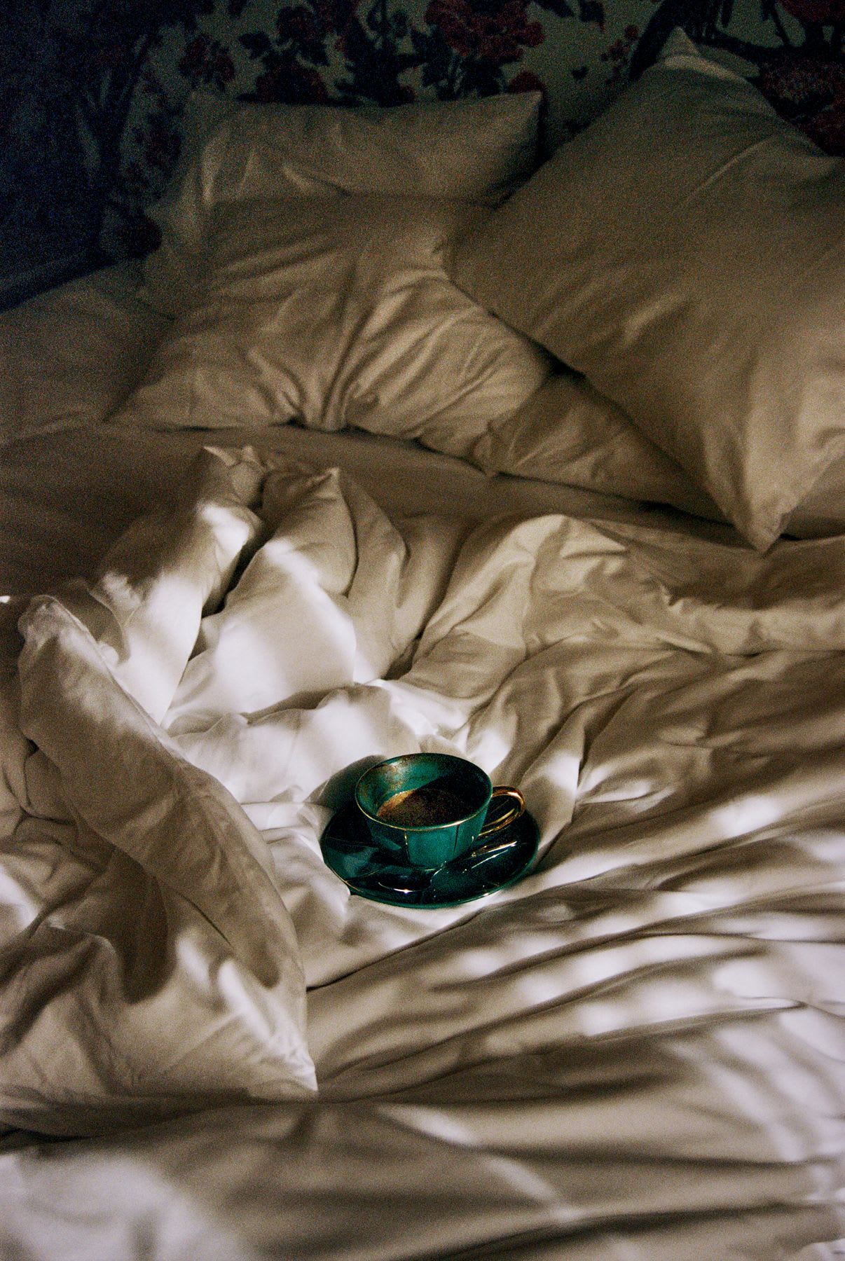An emerald green cup of coffee sits amongst the rumpled white sheets and pillows of an unmade bed.