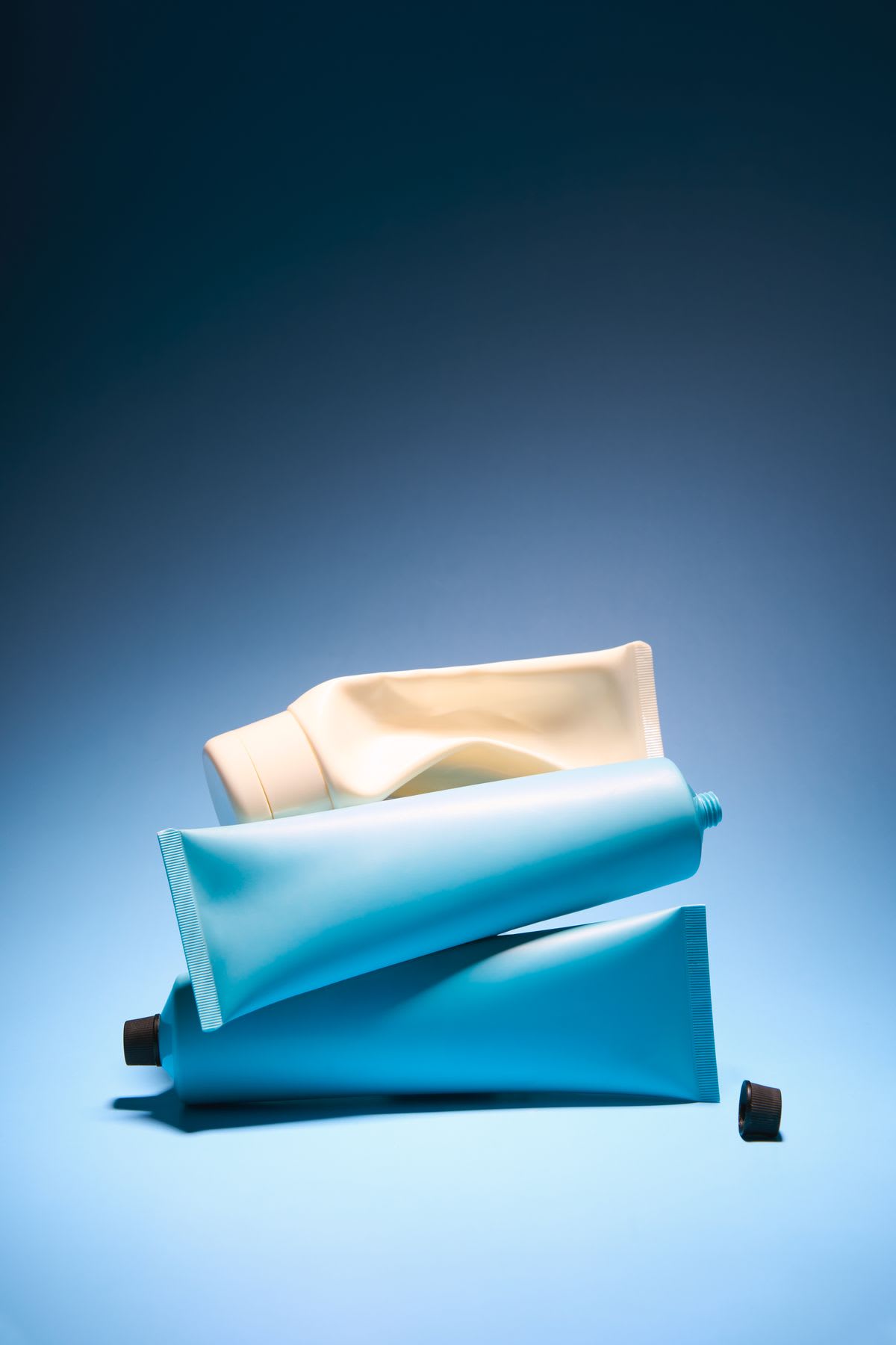 A minimalist still life of two blue cosmetic tubes and one cream-colored tube stacked on a blue gradient background.