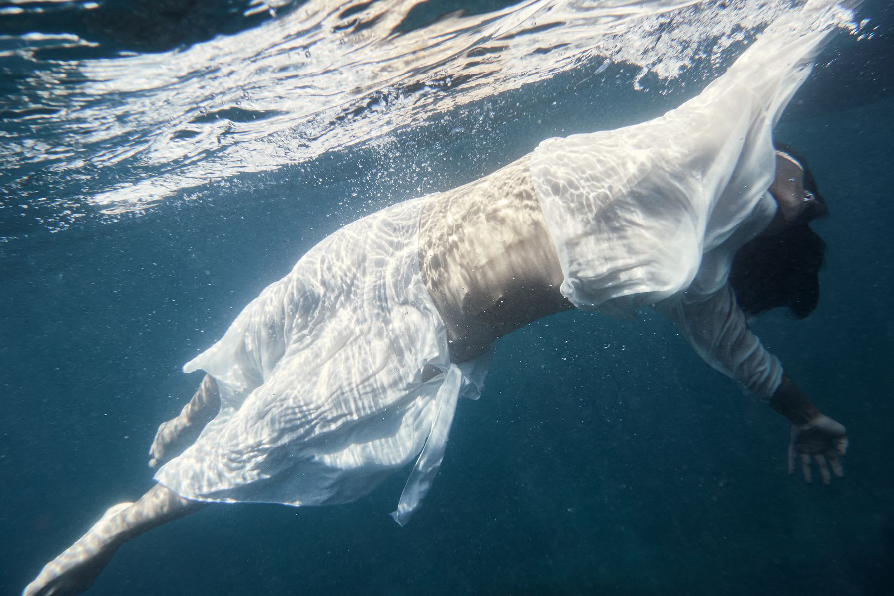 An underwater view of a person wearing a flowing white garment suspended gracefully in deep blue water.