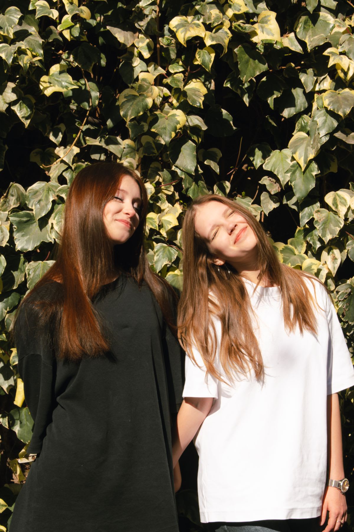 Two girls stand close together smiling in the sun, framed by a wall of ivy leaves in a relaxed outdoor moment.