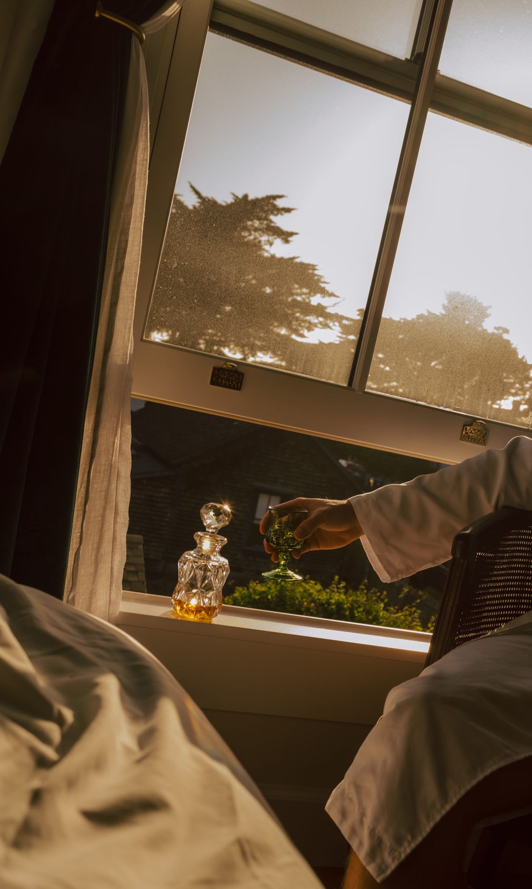 A person wearing a white robe enjoys a drink from a crystal decanter while sitting by a window during golden hour.