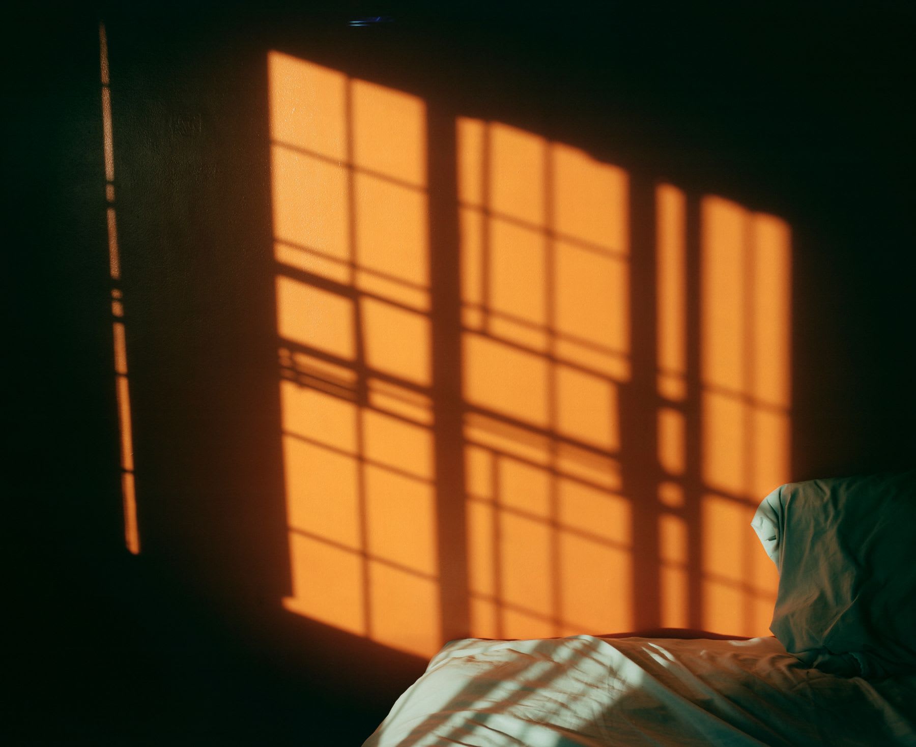 Sunlight streams through a window, casting geometric shadows on the wall and onto an unmade bed.