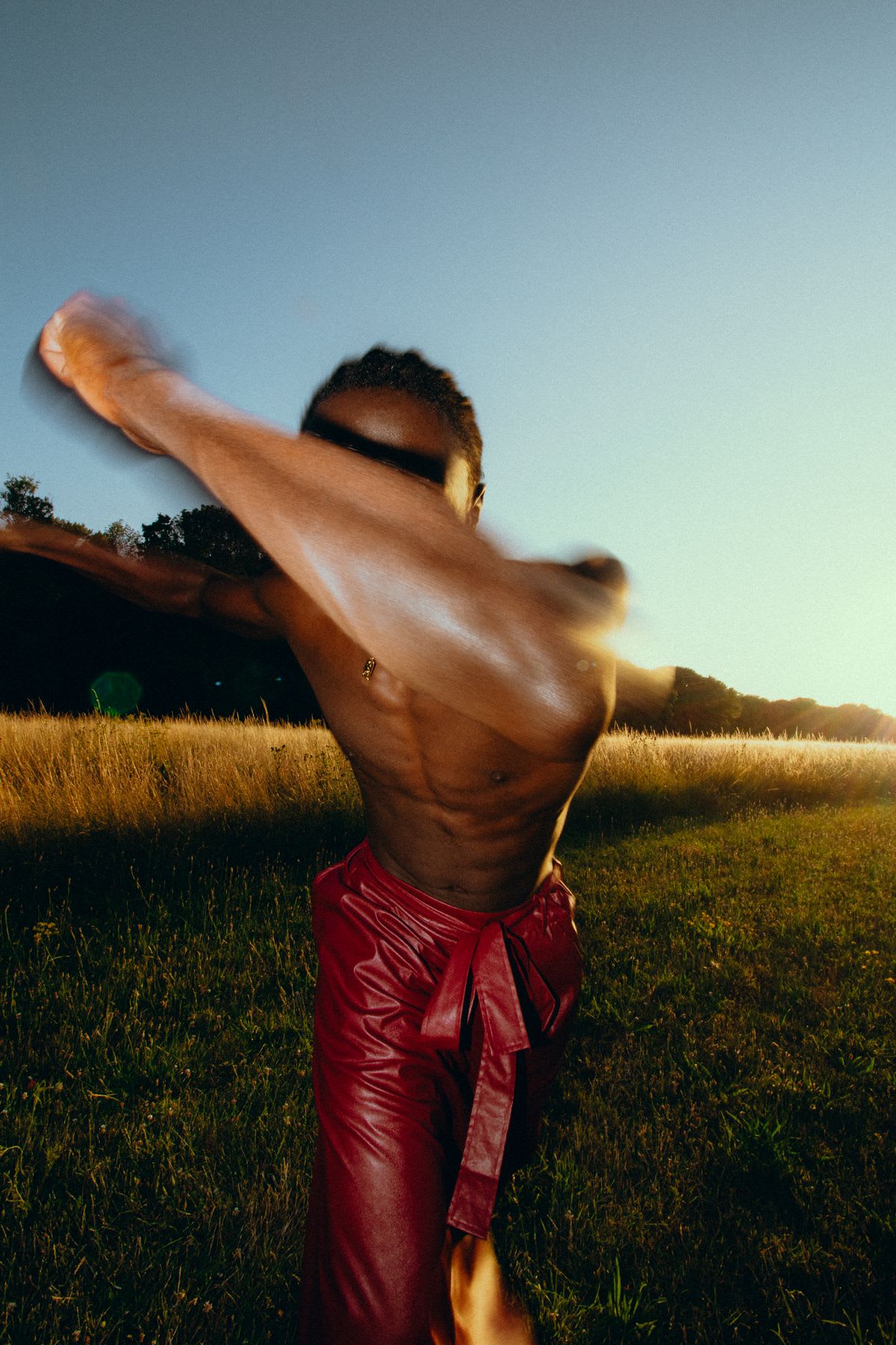 A shirtless young man wearing red trousers walking forward on a grass-covered field at sundown, with his hands up blocking his face.