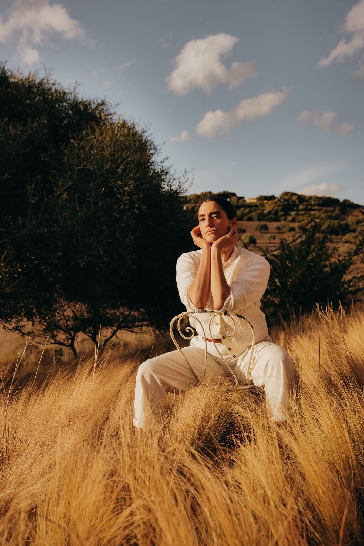 An androgynous model sitting with face in hands on a chair in tall grass.