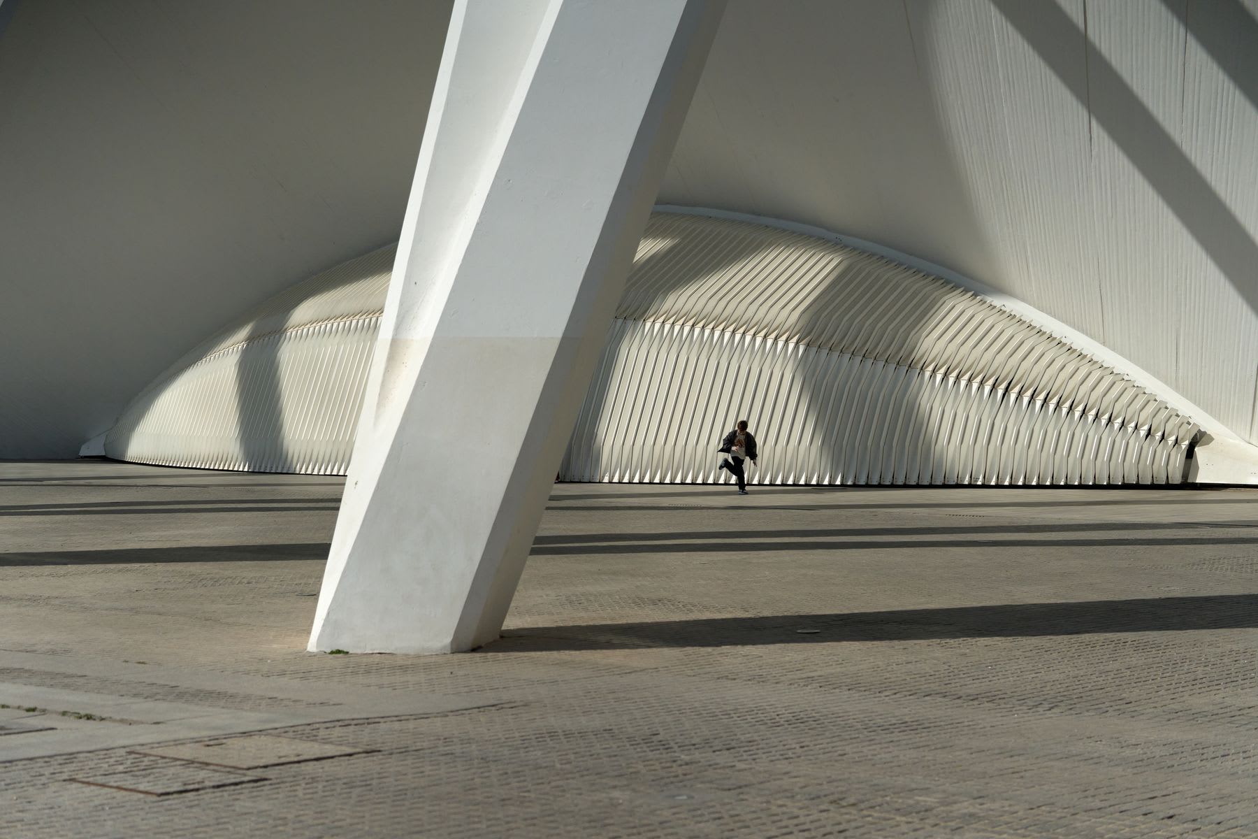 Person runs across the concrete ground behind a giant white support post and in front of a curved wall.