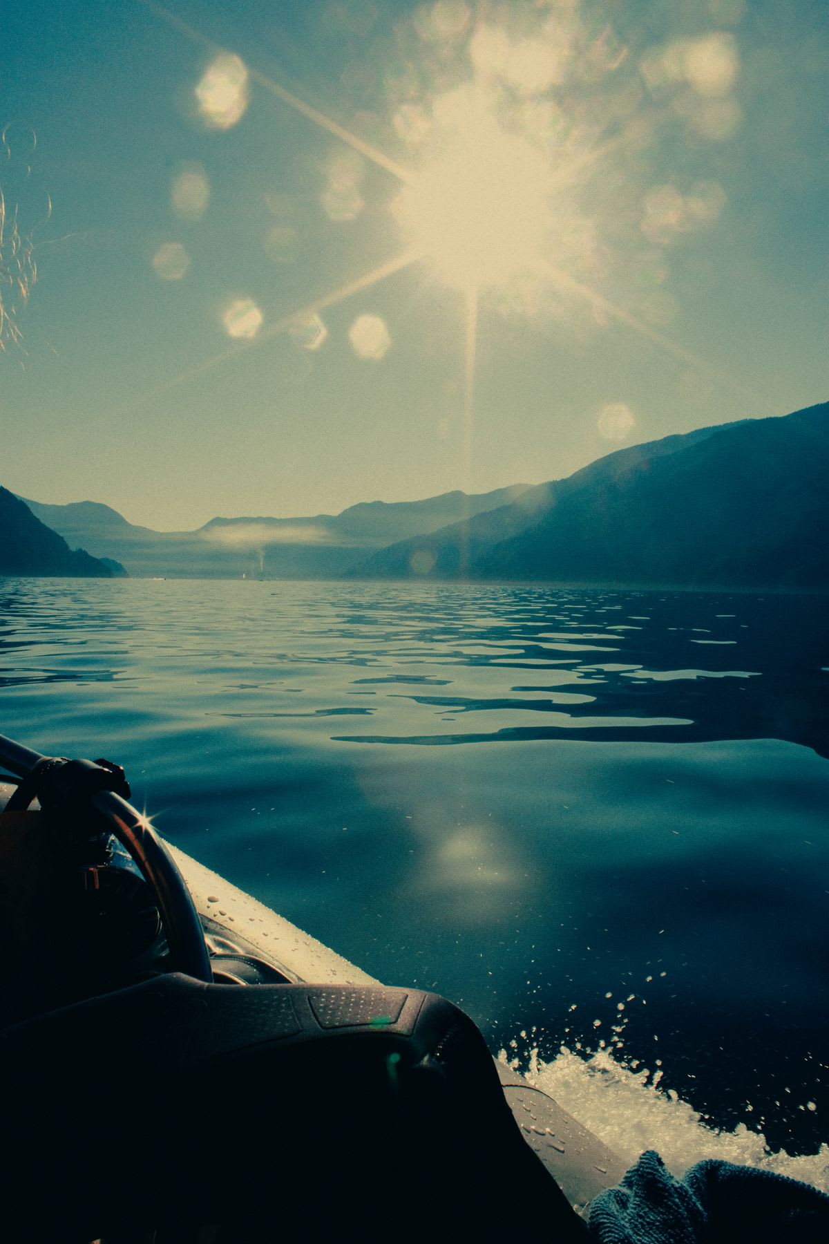 A first-person view from a boat cruising across a tranquil lake surrounded by mountains under the bright morning sun.