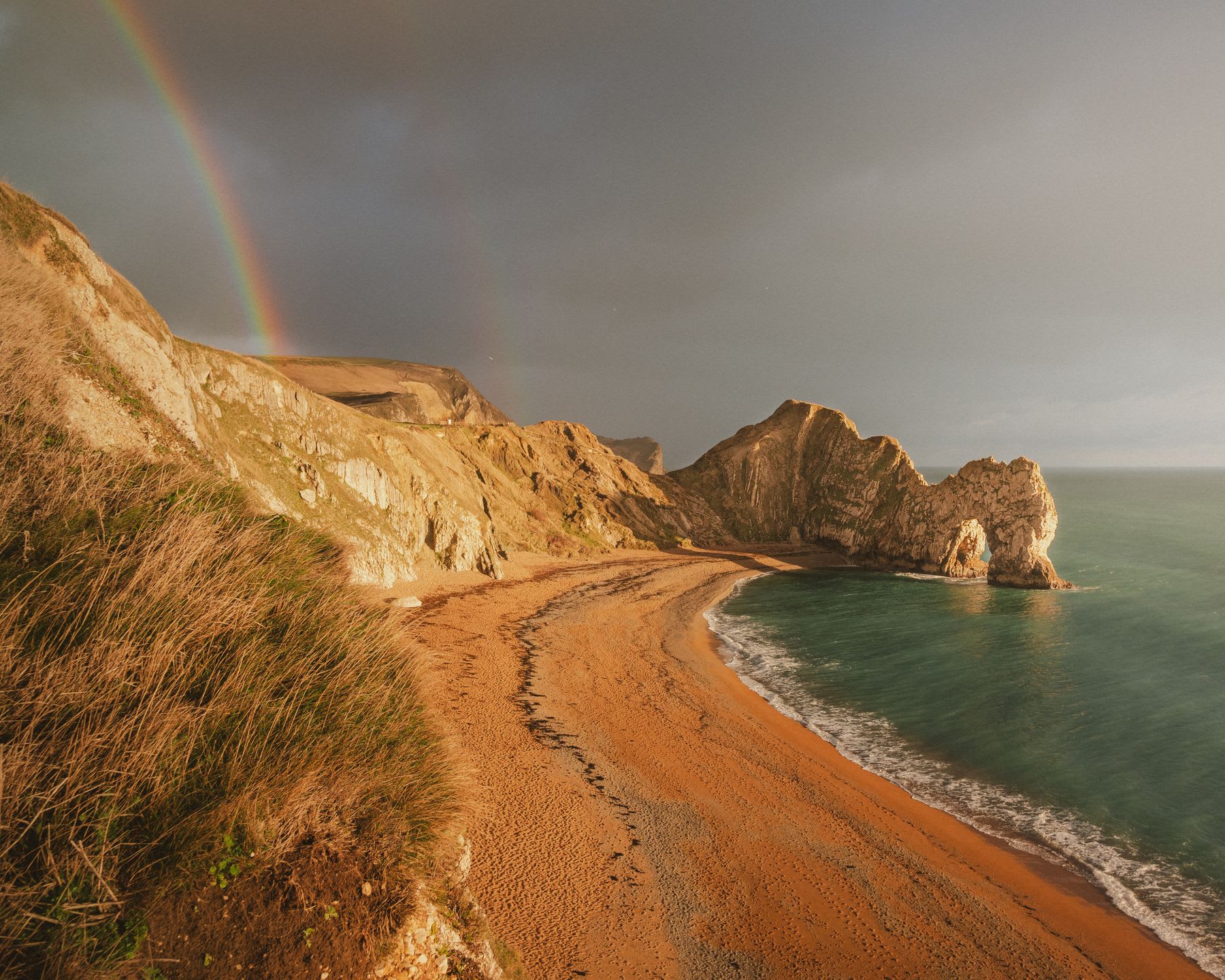 Landscape of a turquoise sea at the edge of the rocky coast on a cloudy day with black clouds and a rainbow