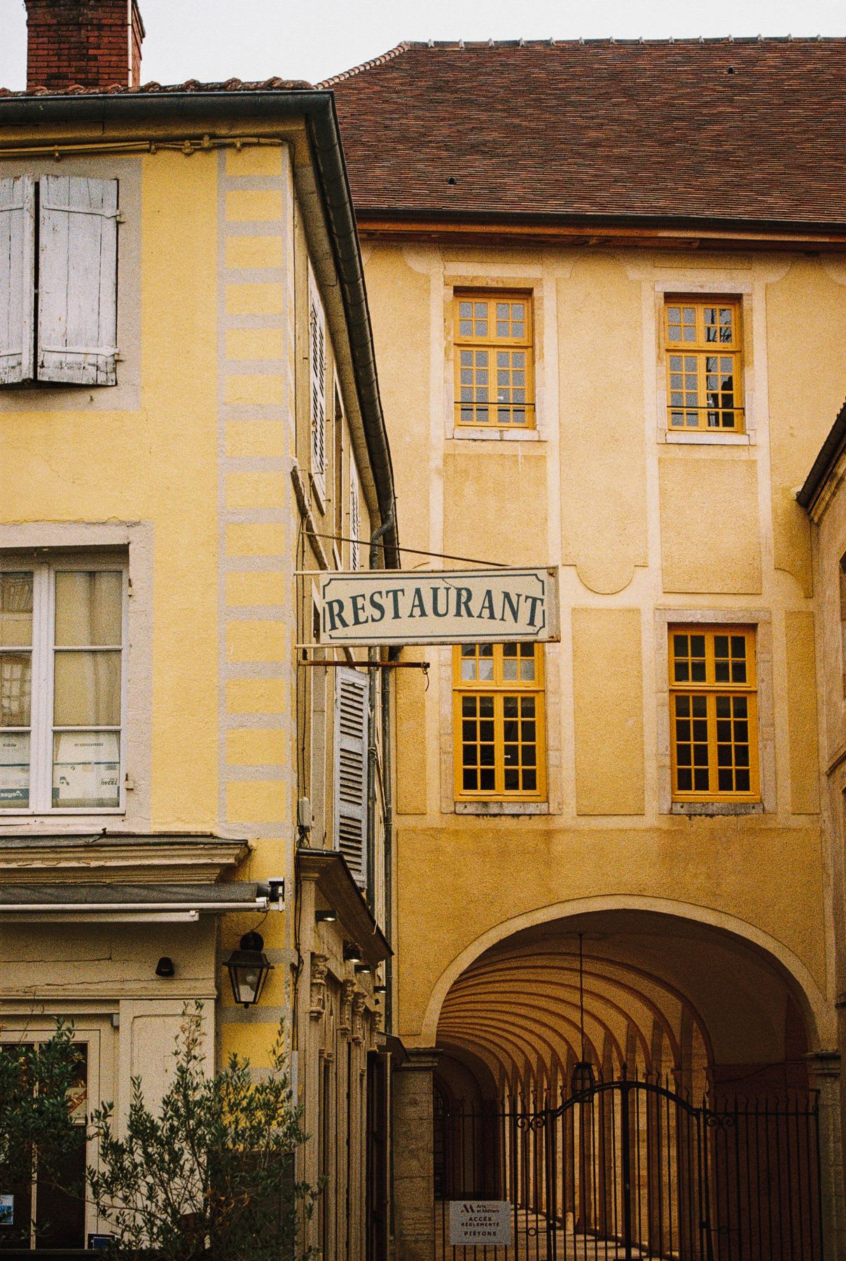 A classic 'Restaurant' sign hangs between two old yellow buildings with an arched entryway in a historic European city.