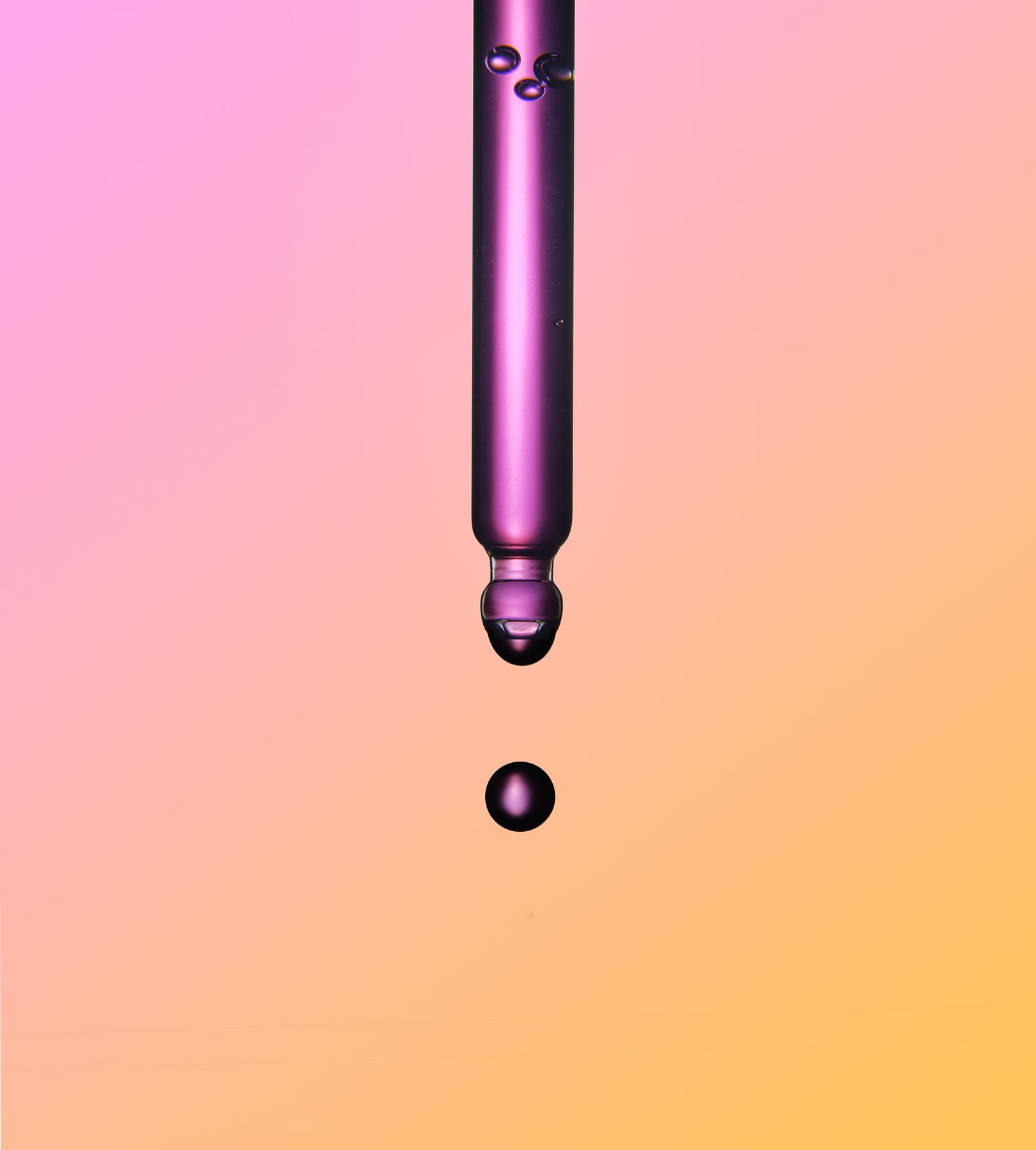 A close-up of a single drop of purple liquid falling from a glass pipette against a pink and yellow gradient background.