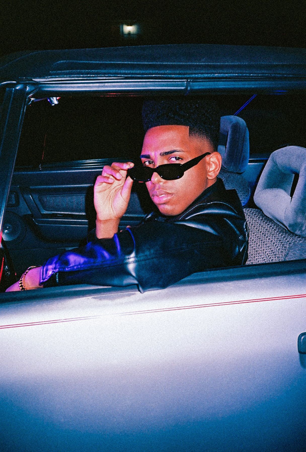 a confident man sitting in a car, wearing a leather jacket and sunglasses, with a bold and stylish vibe illuminated by neon lighting.