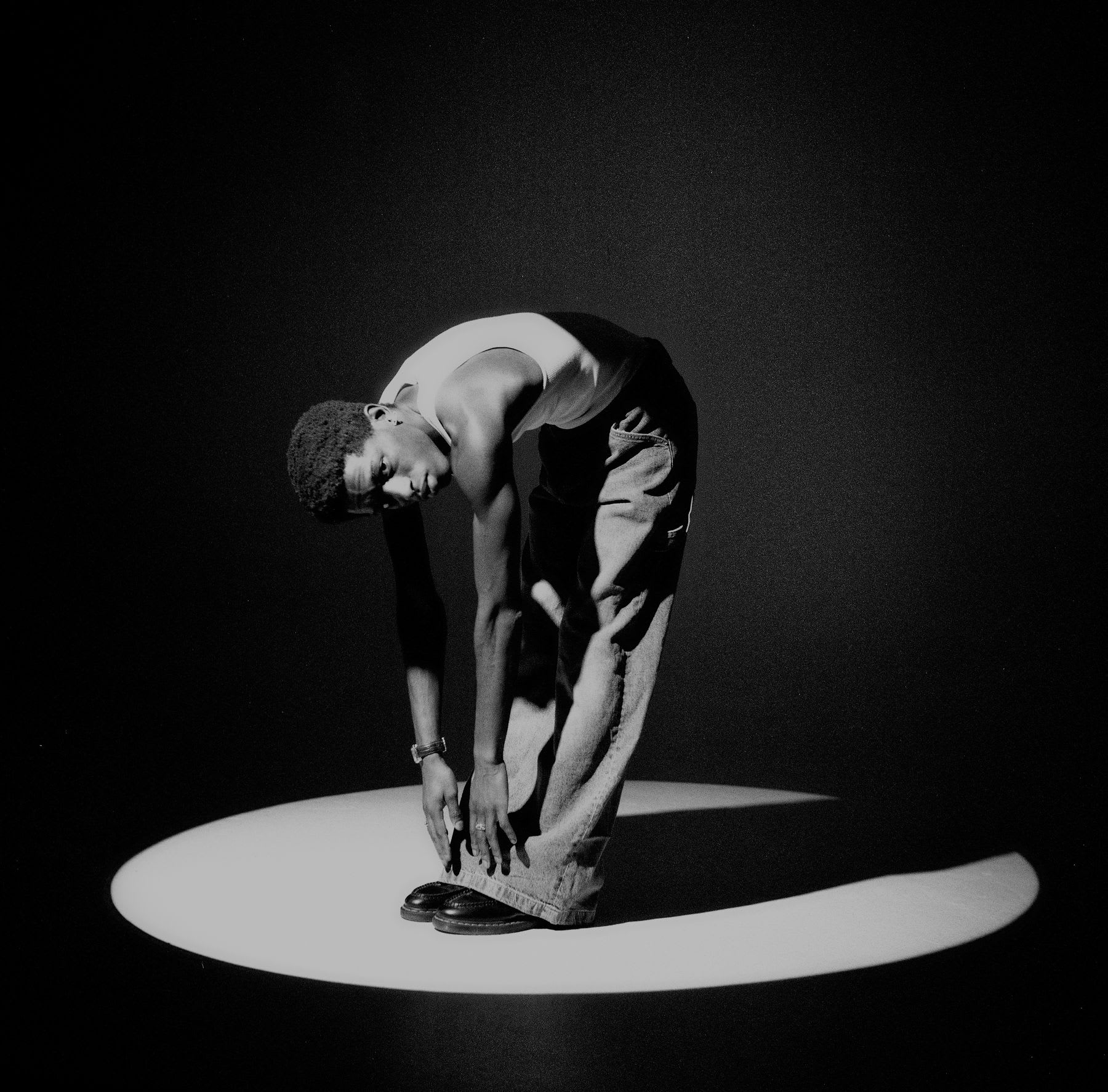 Young man reaches down to his feet while standing in the light cast by a spotlight.