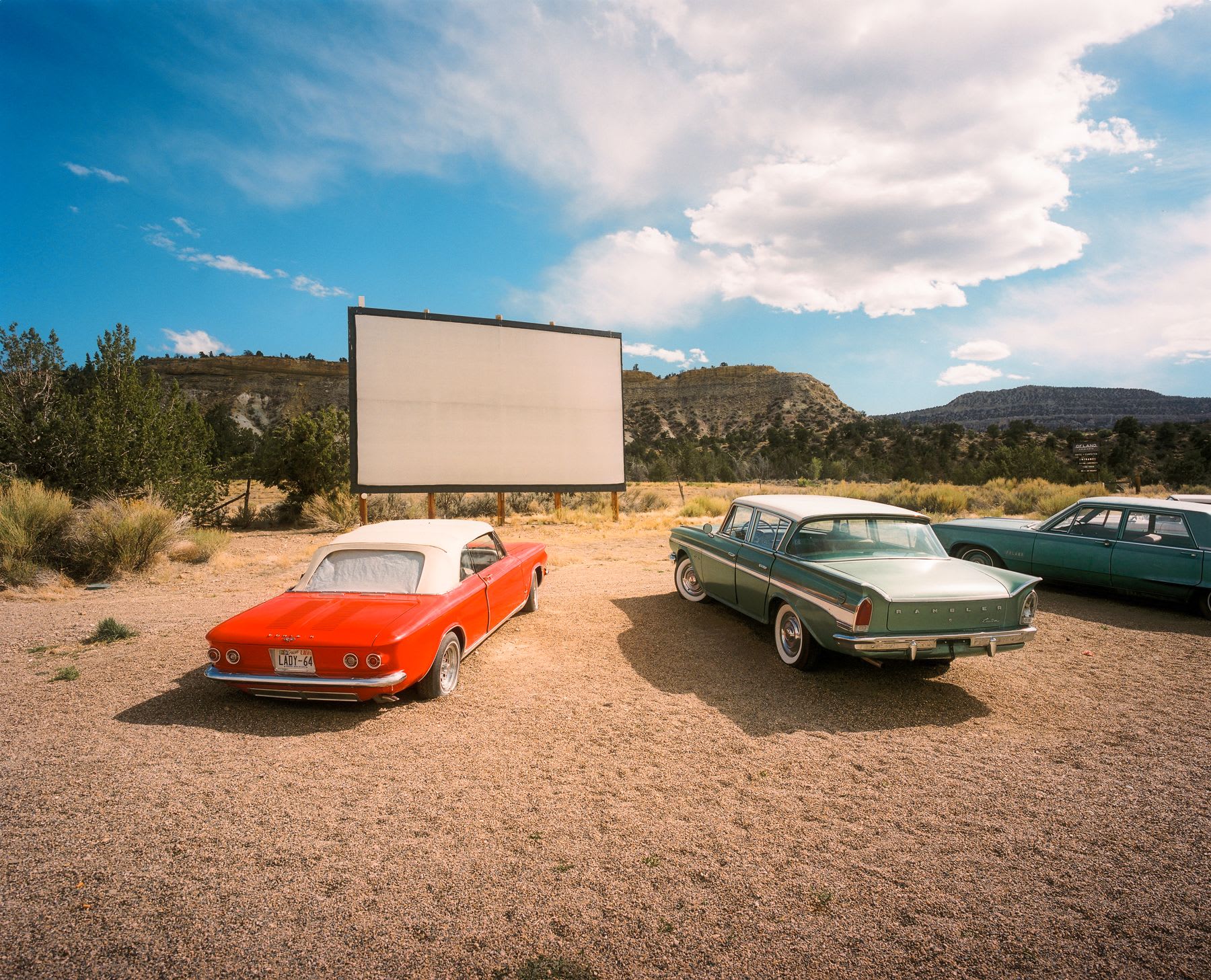 Classic cars are parked on a gravel lot at an outdoor drive-in cinema with mountains in the background.