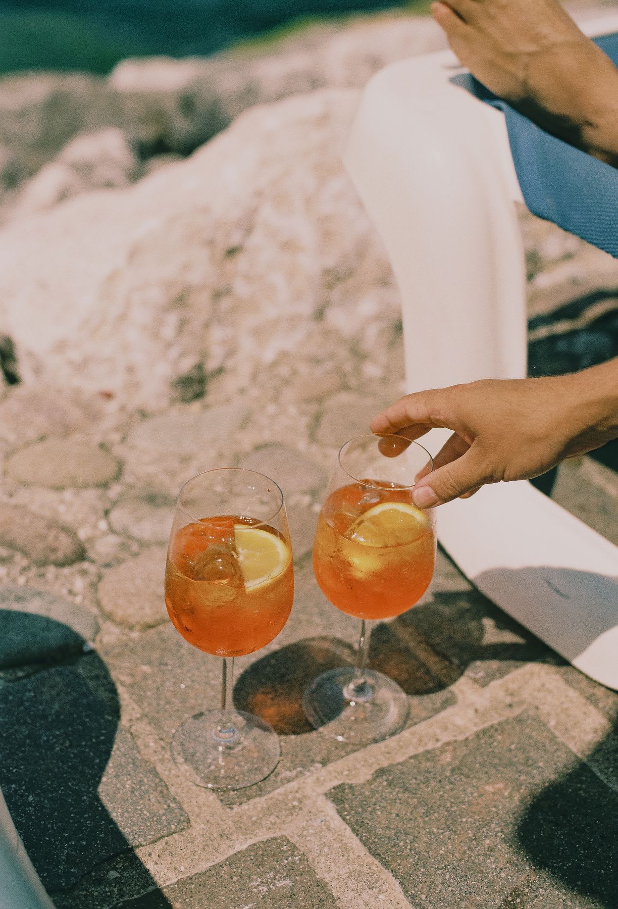 A hand reaches for a glass of a vibrant orange cocktail resting on a sunlit stone patio.
