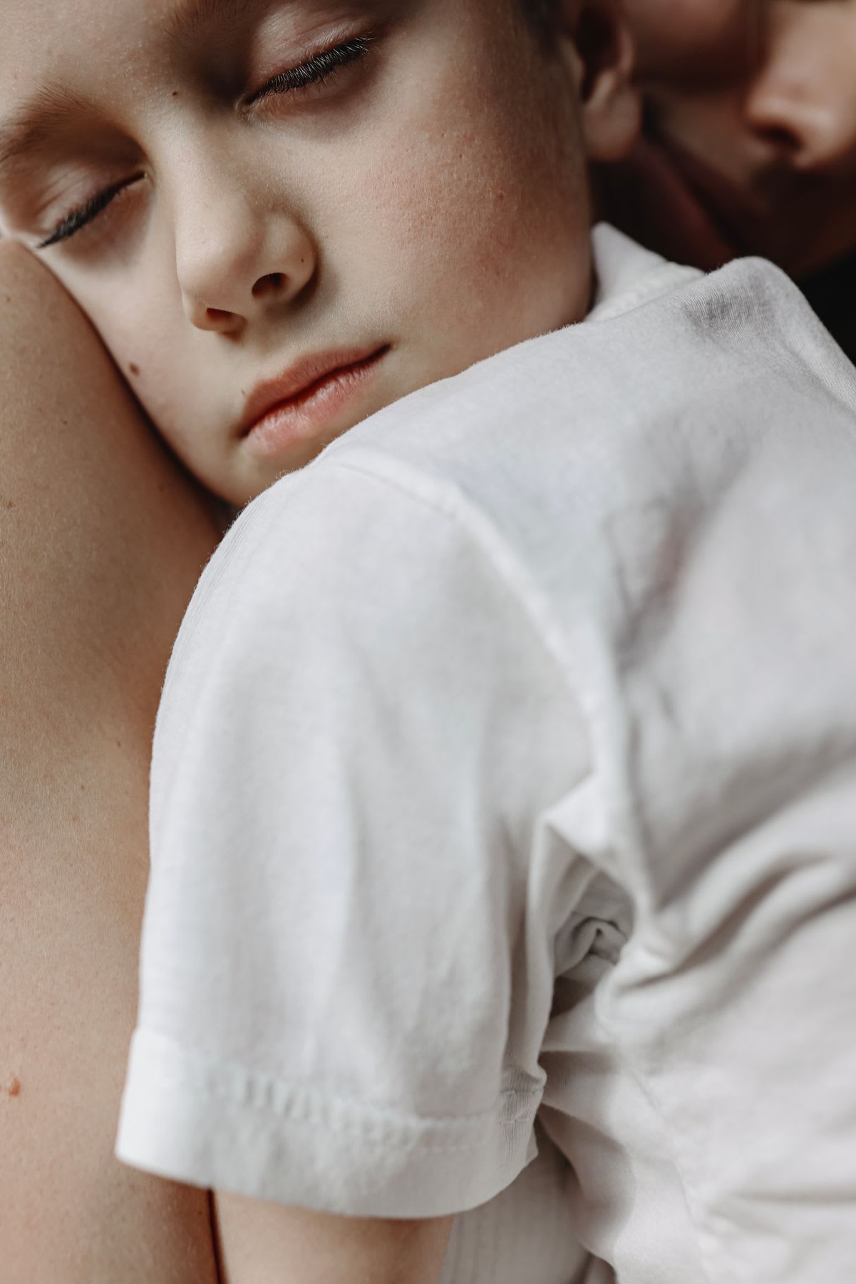 a young child with closed eyes, resting their head on an mother’s shoulder or chest, wearing a white shirt.