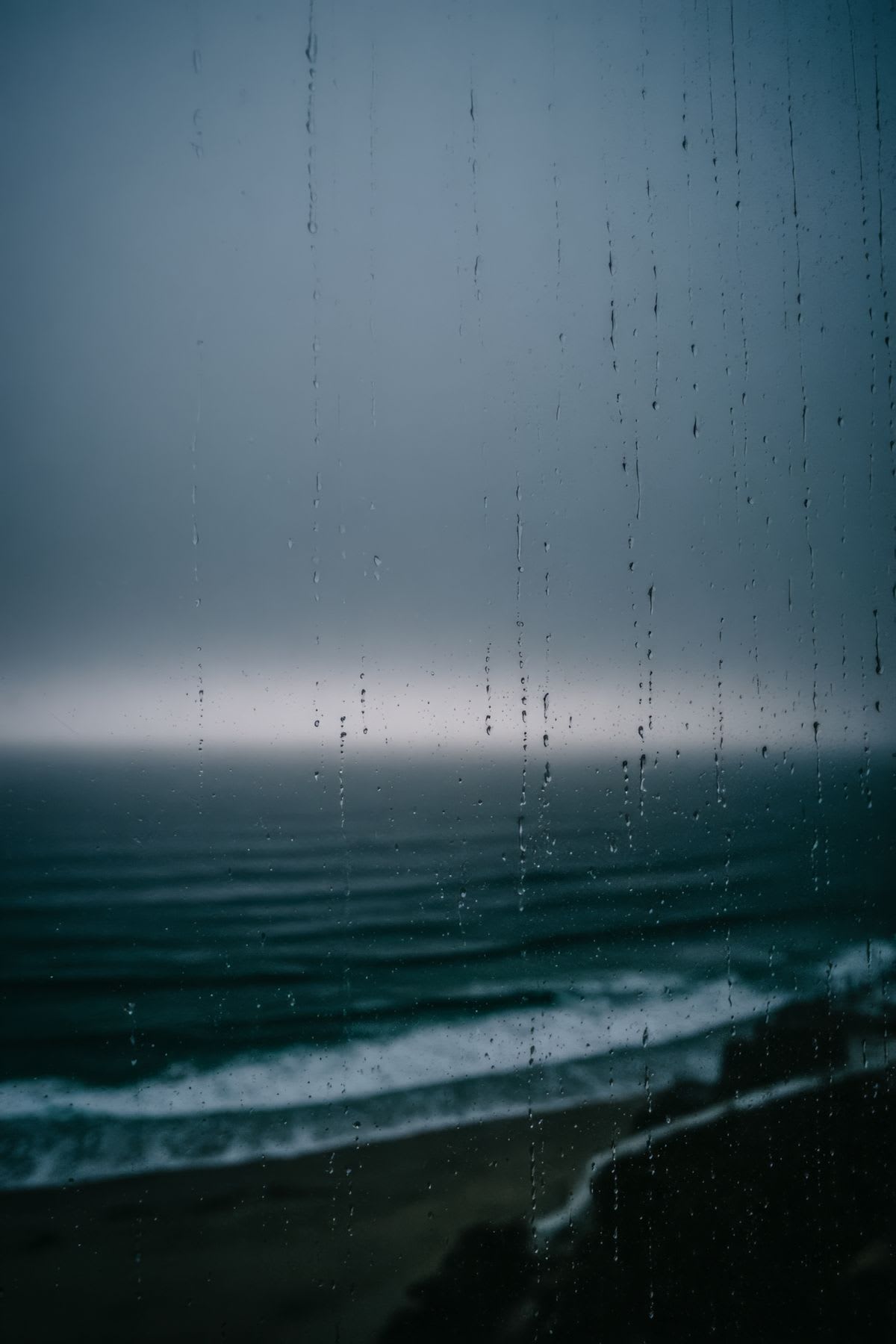 Rain streaks down a window overlooking a somber seascape with dark waves meeting a sandy shore under a heavy sky.
