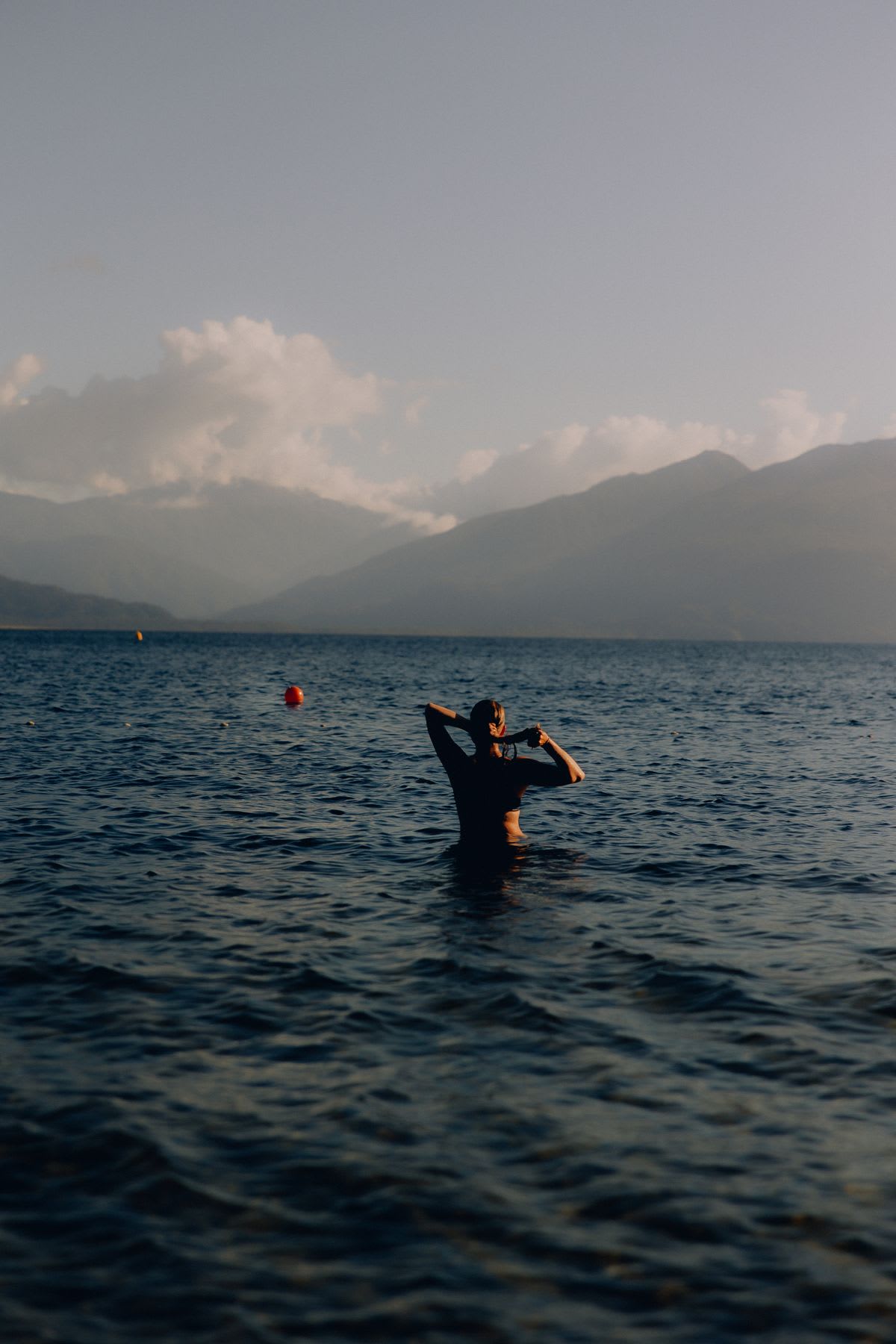 A woman wades into the deep blue water of a mountain lake, preparing for an early morning swim.