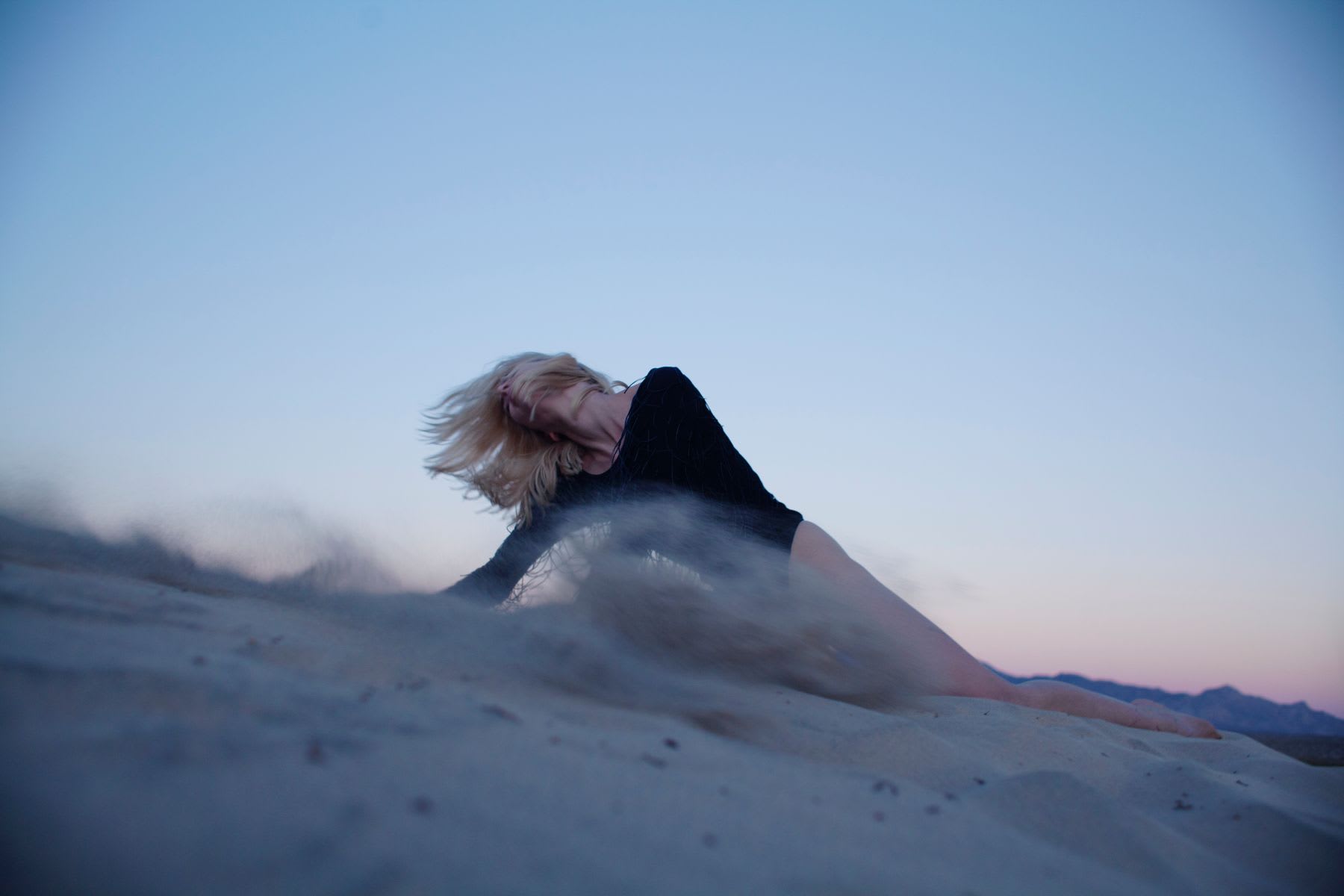 A blonde model with head and hair whipped back posed on a sand dune at sunset