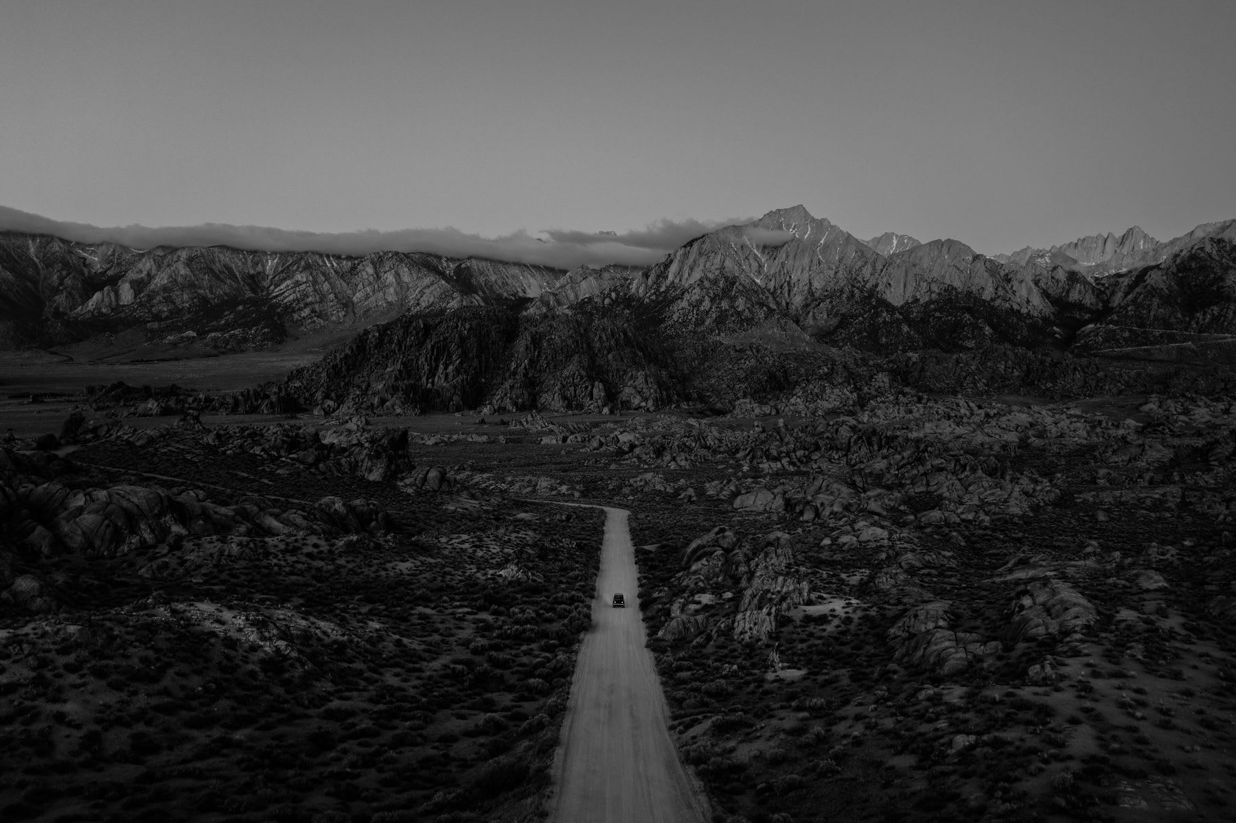 A road with a single car in front of a mountain range in black and white